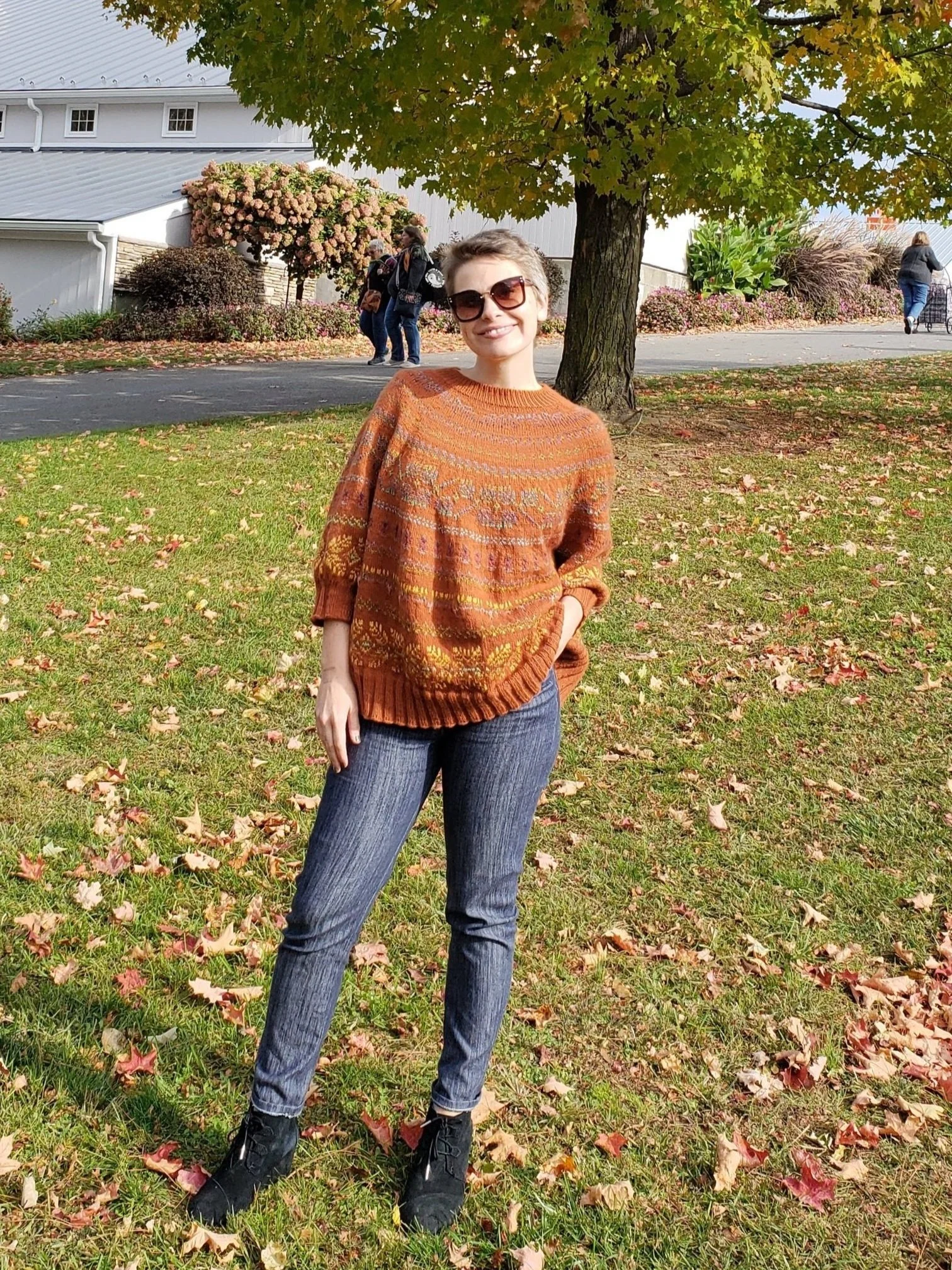 A woman standing outdoors on a grassy area with fallen autumn leaves, wearing sunglasses, a patterned orange sweater, jeans, and black boots, smiling.