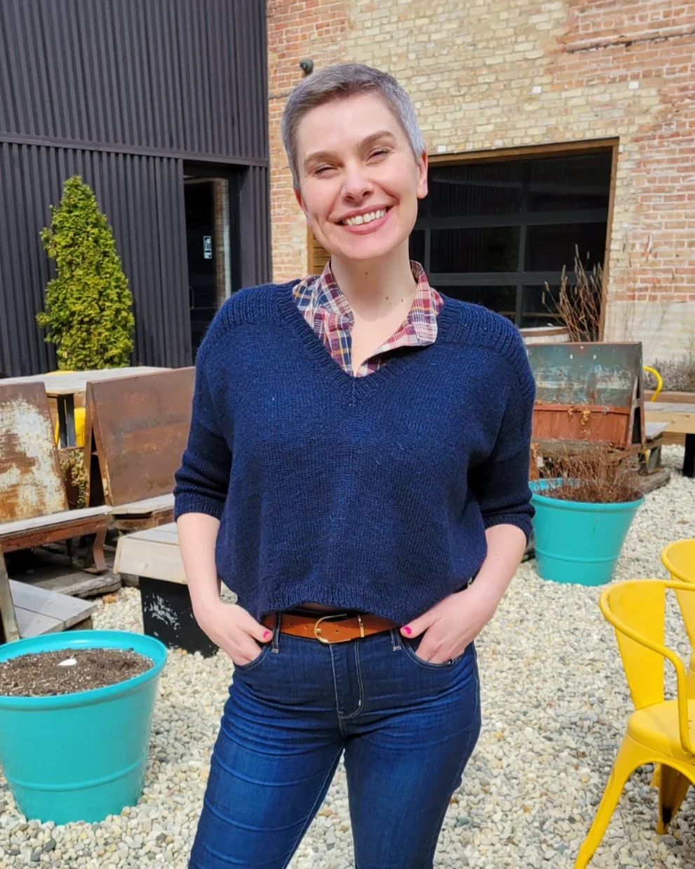 A woman with short gray hair wearing a blue sweater, plaid shirt, and jeans standing outdoors in front of colorful chairs and planters, smiling with her hands in her pockets.