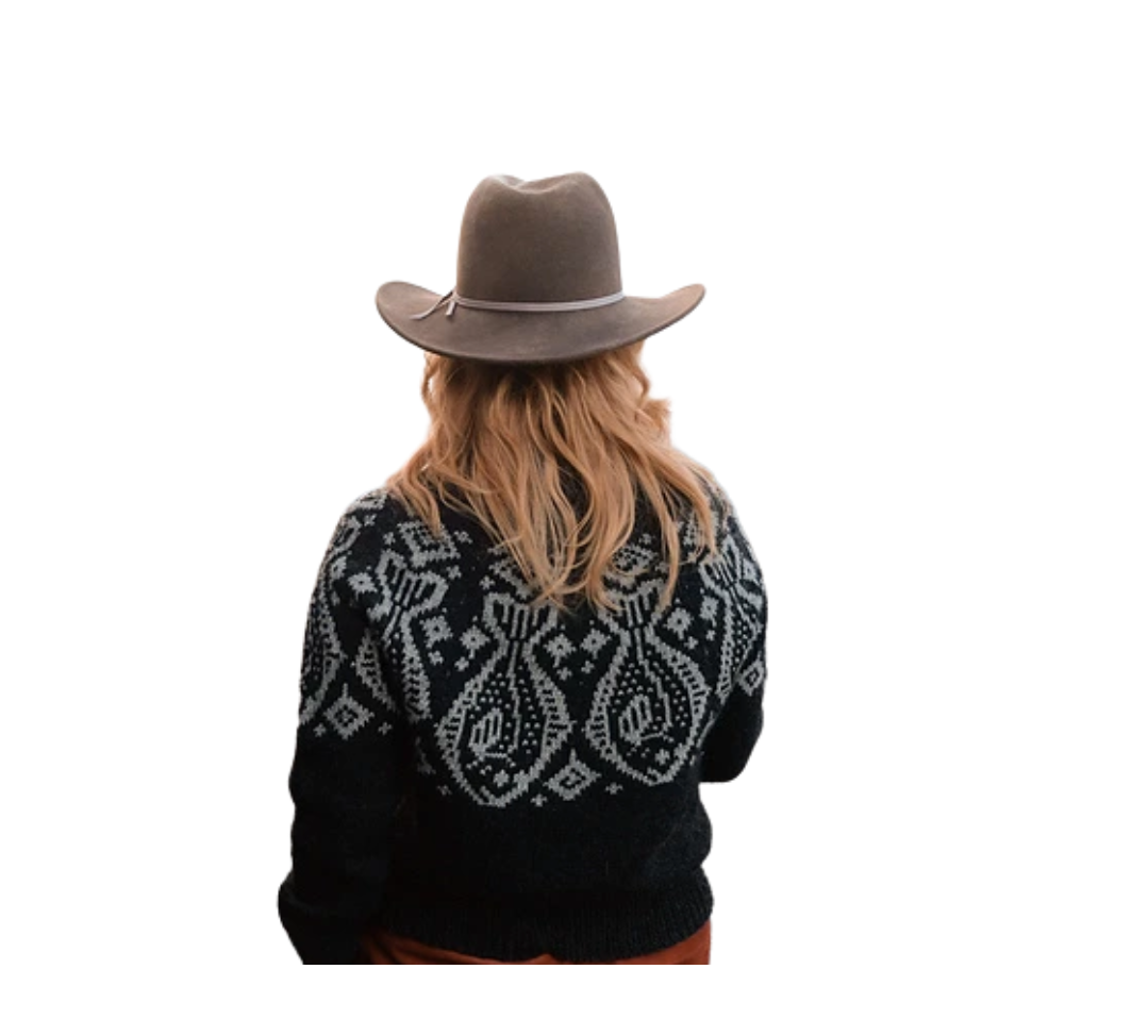 Back view of a woman with wavy red hair wearing a wide-brimmed gray hat and a black sweater with a white Nordic pattern.