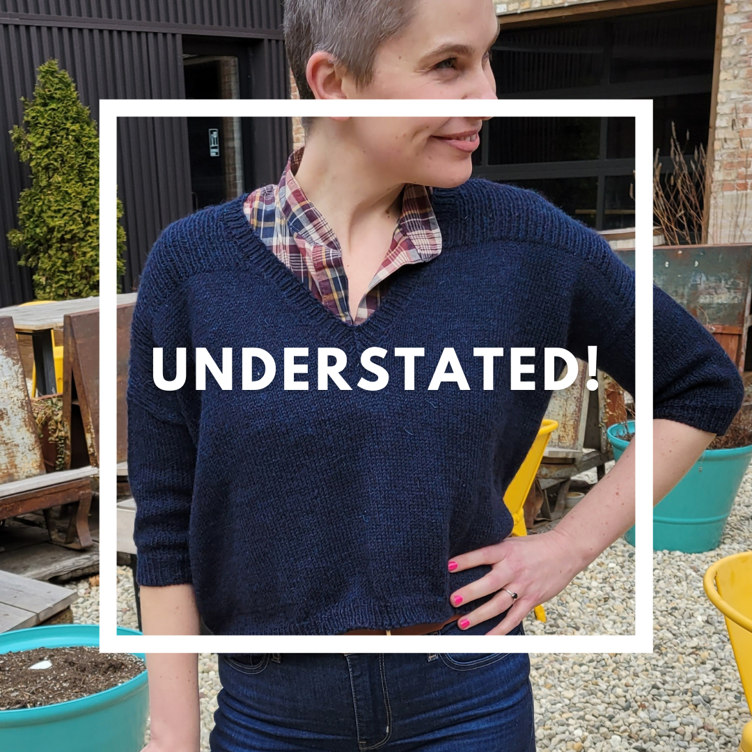 A woman smiling outdoors, wearing a plaid shirt under a dark blue sweater. She has short hair and is standing with her hand on her hip, with colorful plant pots and outdoor furniture in the background.
