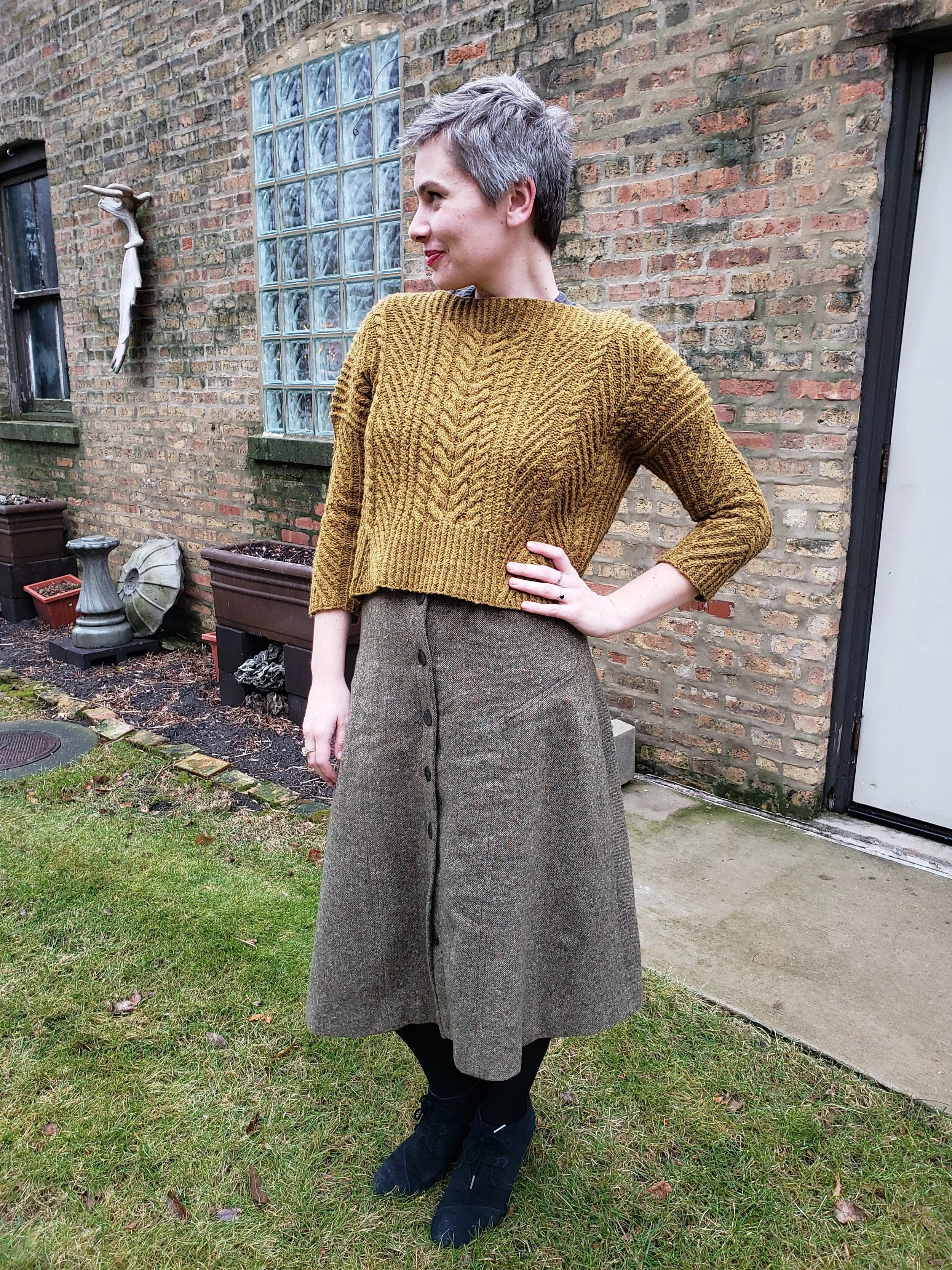 Woman with short gray hair wearing a mustard yellow sweater, brown button-front skirt, black tights, and black shoes standing outdoors on grass near a brick wall with potted plants and a window.