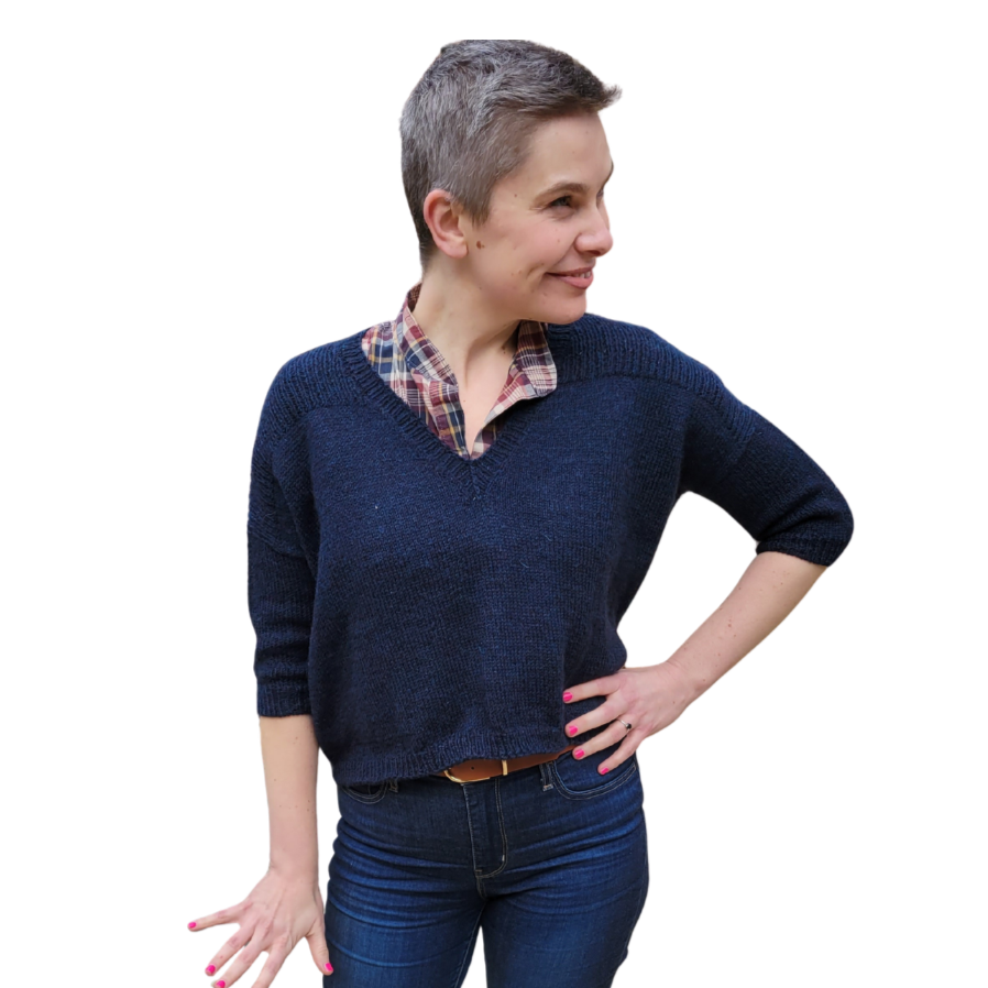 A woman with short gray hair wearing a navy blue sweater over a plaid shirt, standing with one hand on her hip and smiling, against a plain white background.