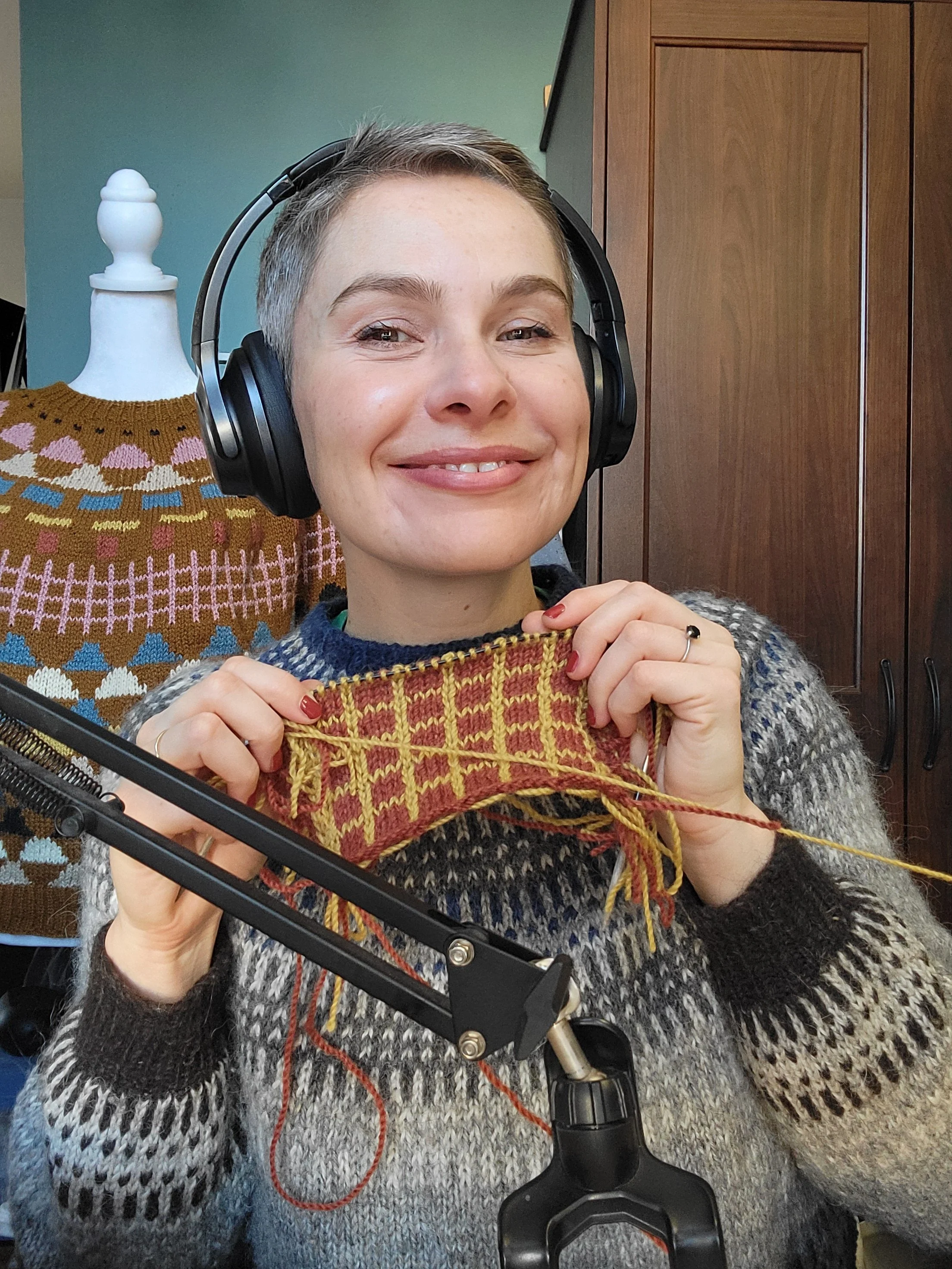 A woman with short hair wearing headphones and a multicolored sweater, smiling and holding a knitted piece of fabric in front of her, with a colorful sweater display behind her.