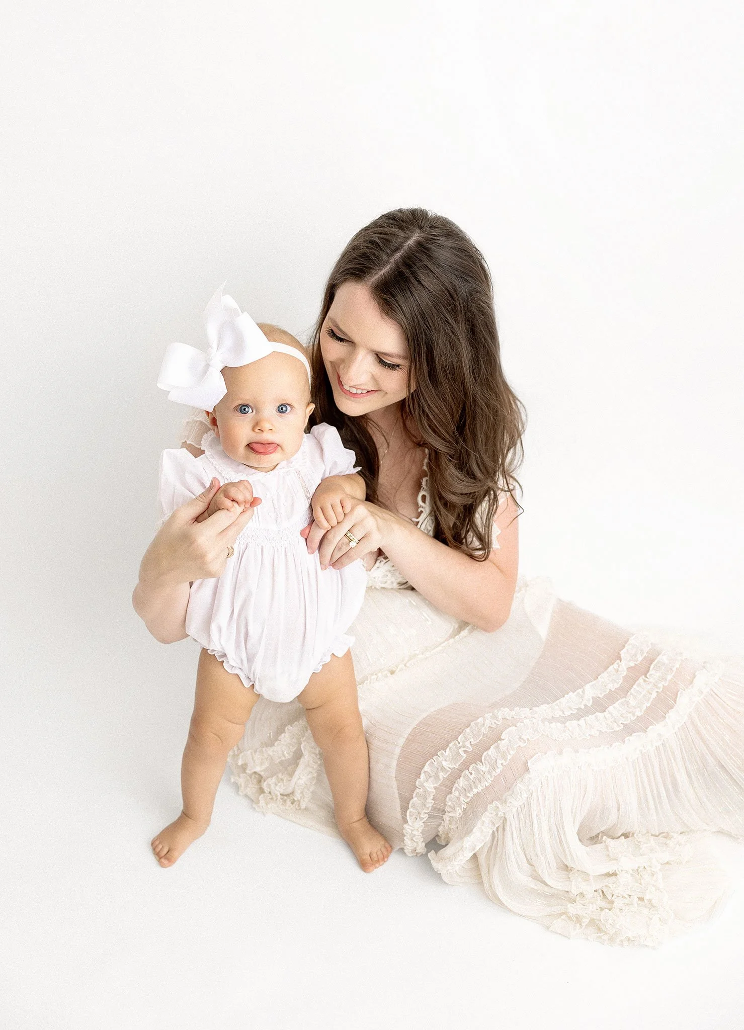 Woman sitting on the floor holding a smiling baby wearing a white dress and headband.