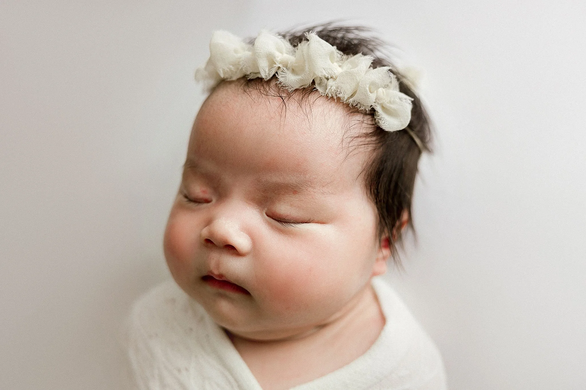 Close-up of a sleeping baby wearing a cream-colored headband with fabric flowers, against a plain white background.