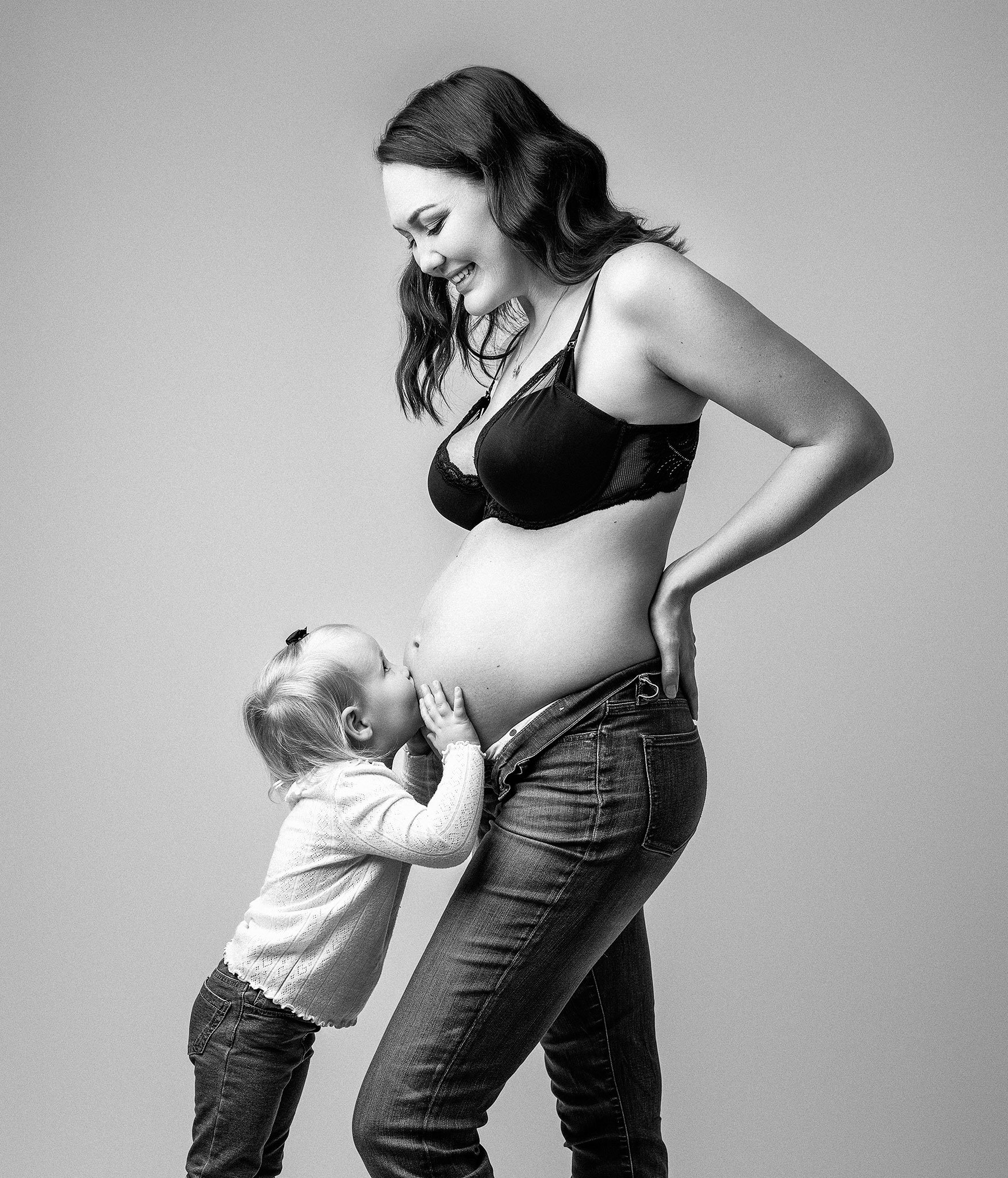 Pregnant woman standing in side profile wearing a bra and jeans, with a young child kissing her pregnant belly. Black and white portrait.