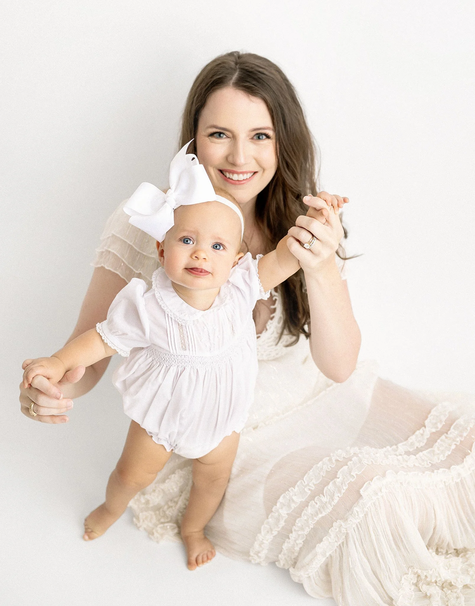 A smiling woman in a white dress holds her baby who is dressed in white with a large bow on her head. The background is plain white, emphasizing the subjects.