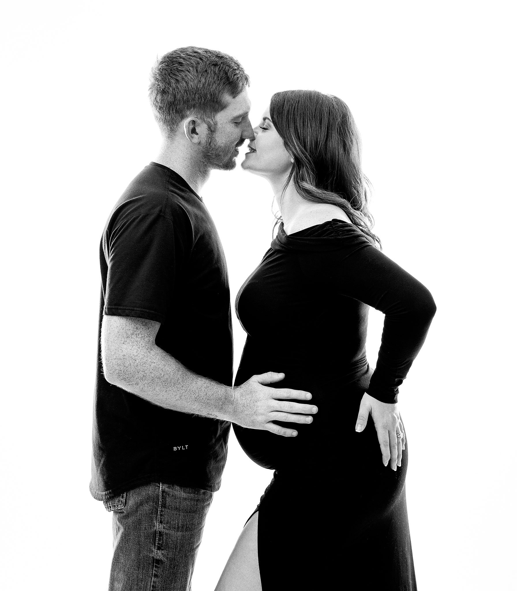 Pregnant woman in black dress kissing a man in a black T-shirt and jeans, both facing each other in a studio setting.