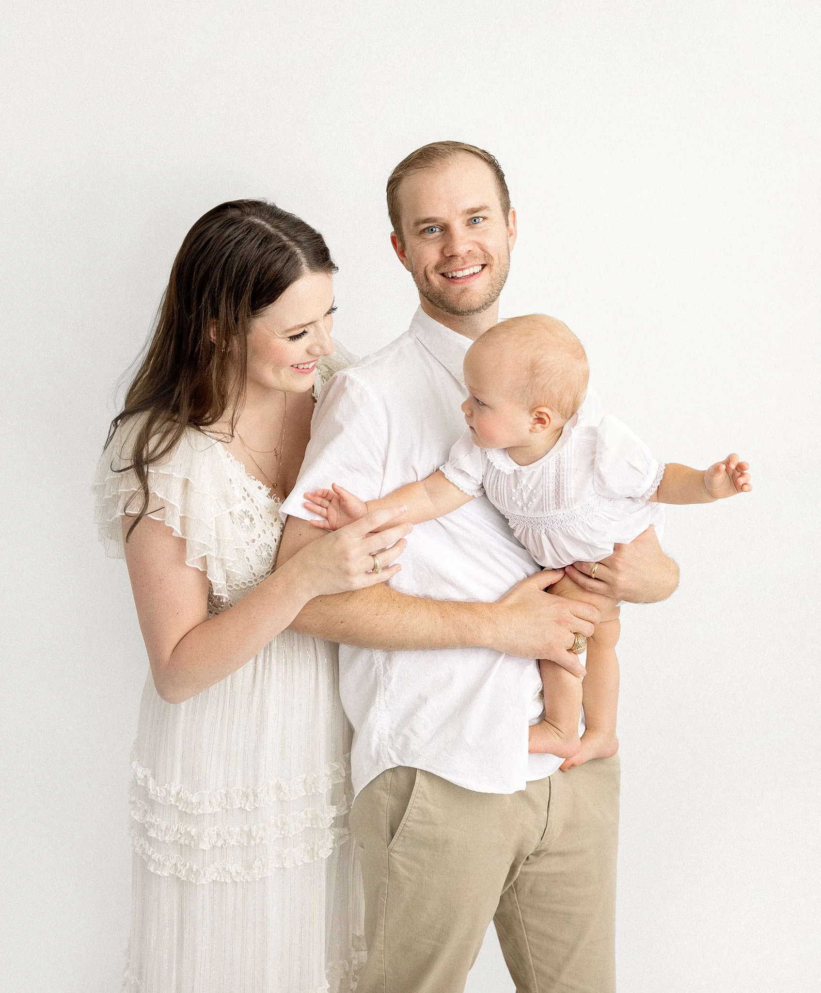 Happy family portrait with a man, woman, and baby in white clothing against a white background.