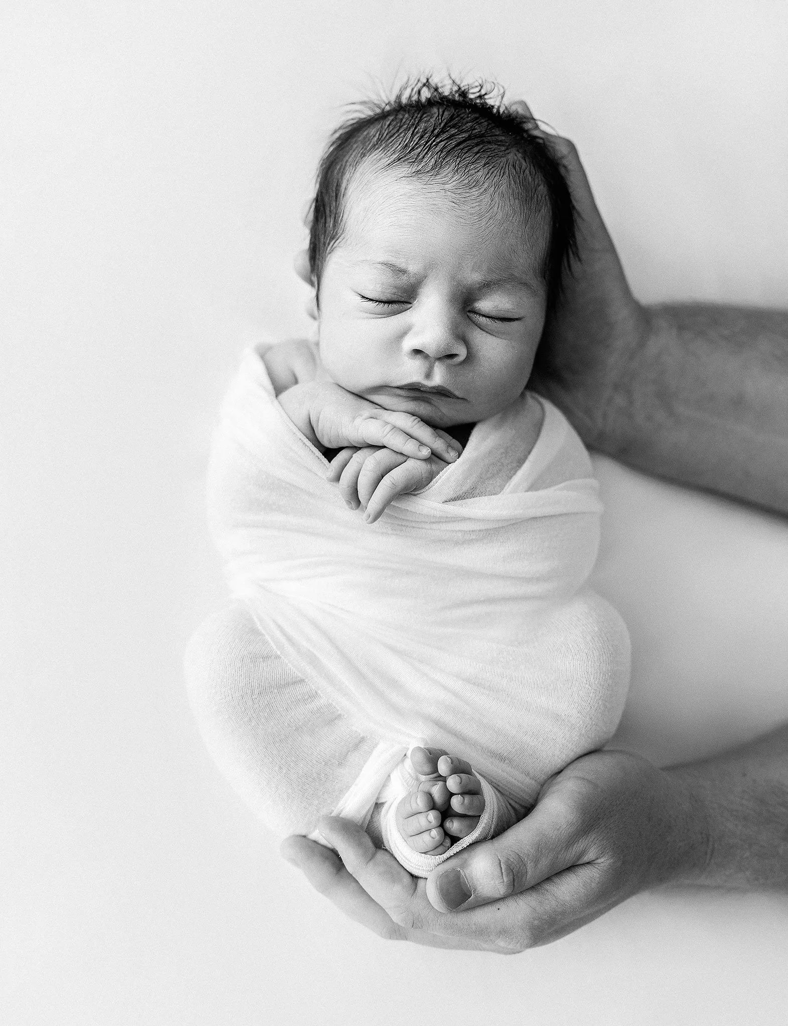 Black and white photo of a newborn baby wrapped in a blanket, held in an adult's hands, eyes closed.