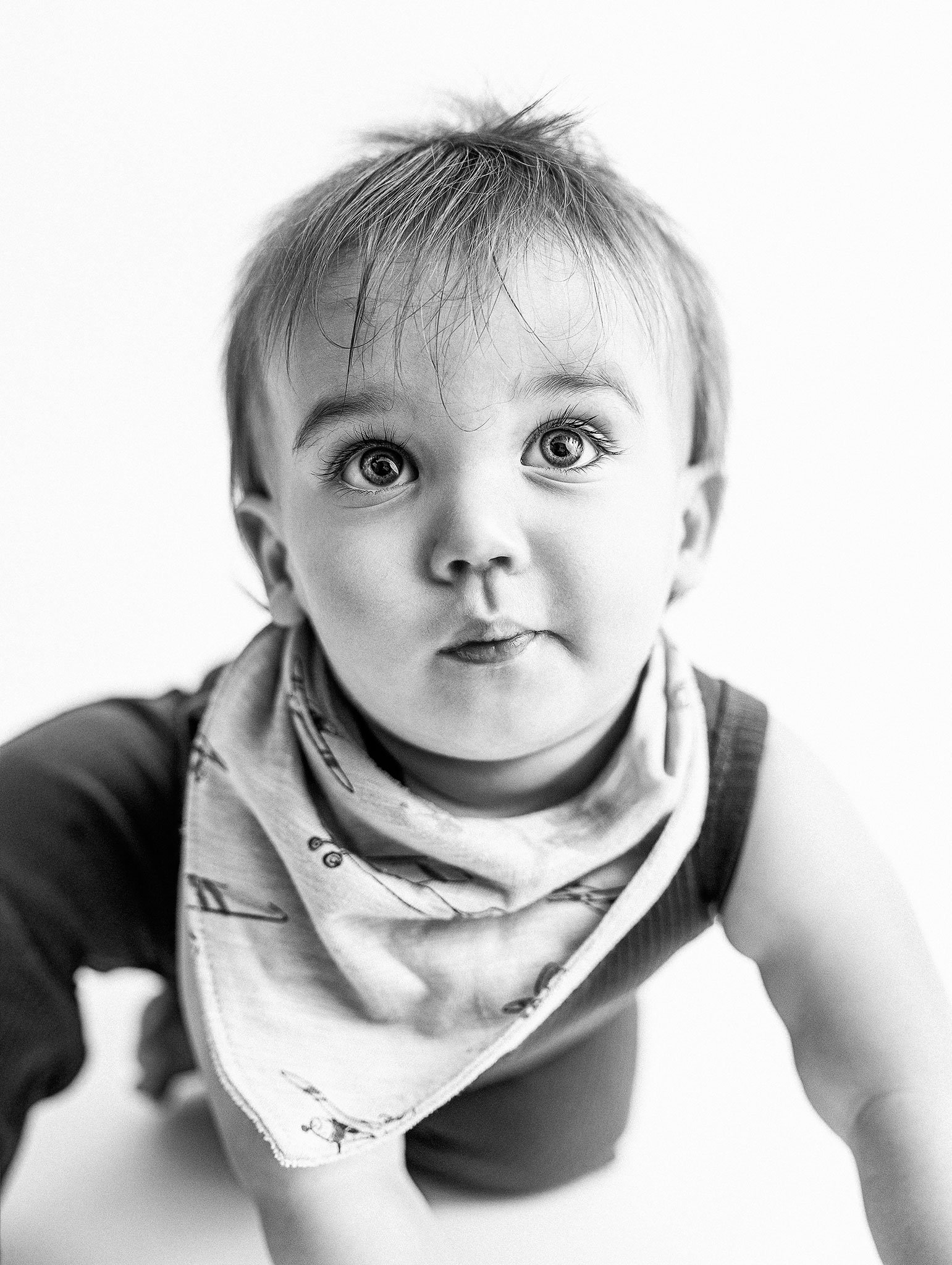 Black and white photo of a baby with wide eyes, wearing a bib with a pattern and sleeveless top, looking at the camera.