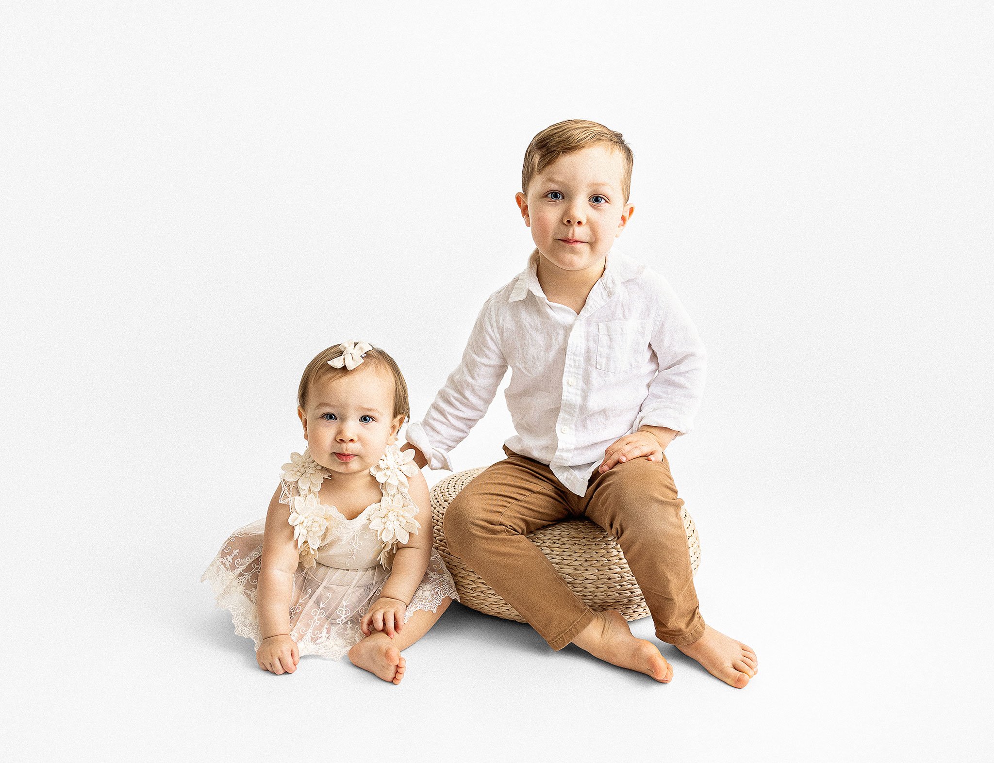 Young boy and girl in light clothing, sitting and smiling on a white background.
