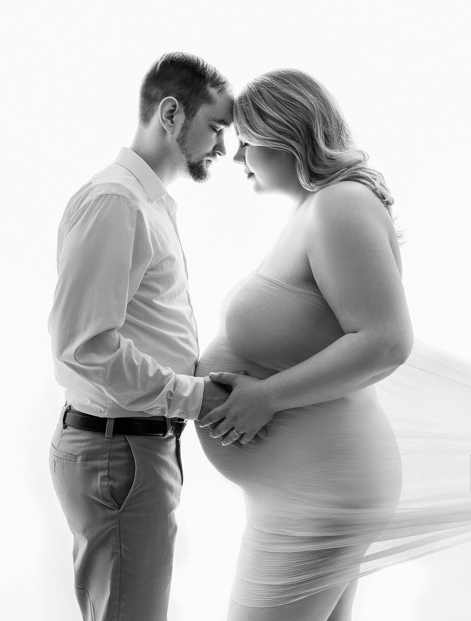 A black and white photograph of a couple standing closely, with foreheads touching. The woman, wearing a flowing dress, is pregnant and both are touching her belly.
