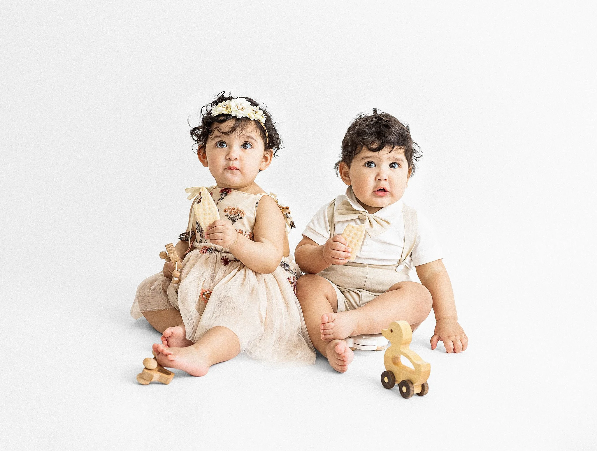 Two young children, a girl and a boy, sitting on the floor against a white background, holding wooden toys and eating snacks, dressed in light-colored clothing.