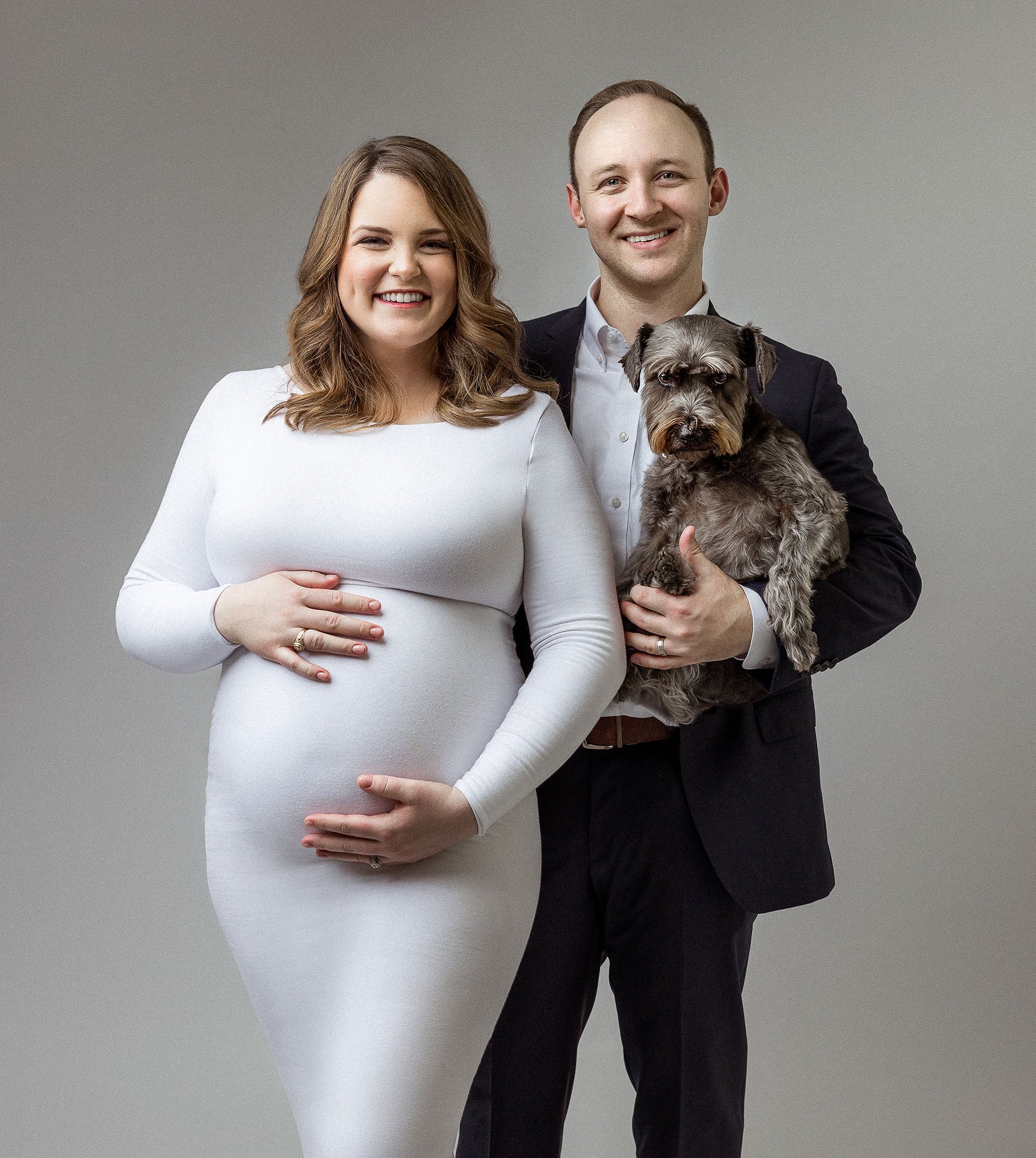 Pregnant woman in a white dress standing with a man in a suit holding a small dog.