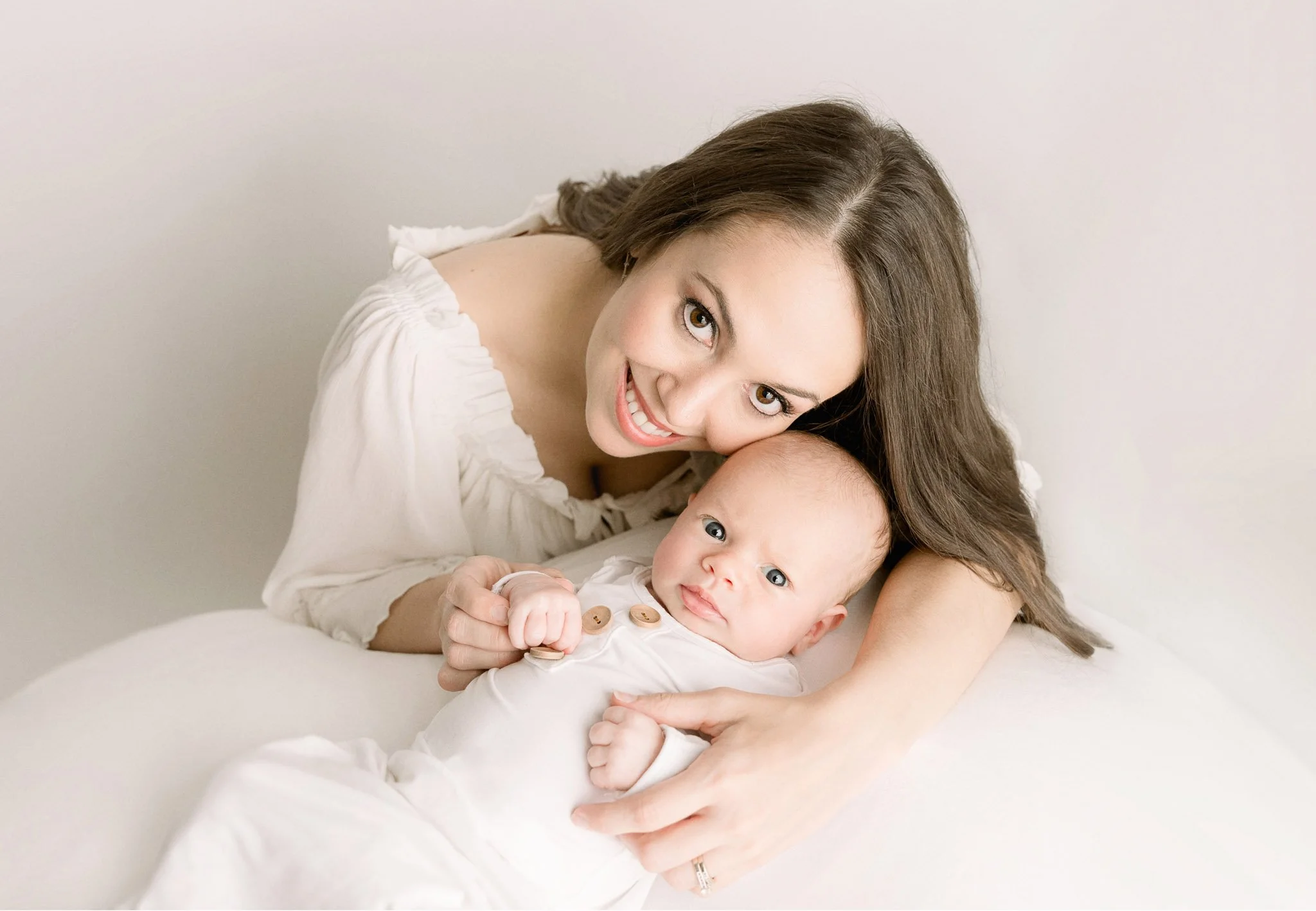 A woman smiling and lying next to a baby dressed in a white outfit, both on a white background. The woman is holding the baby's hand.