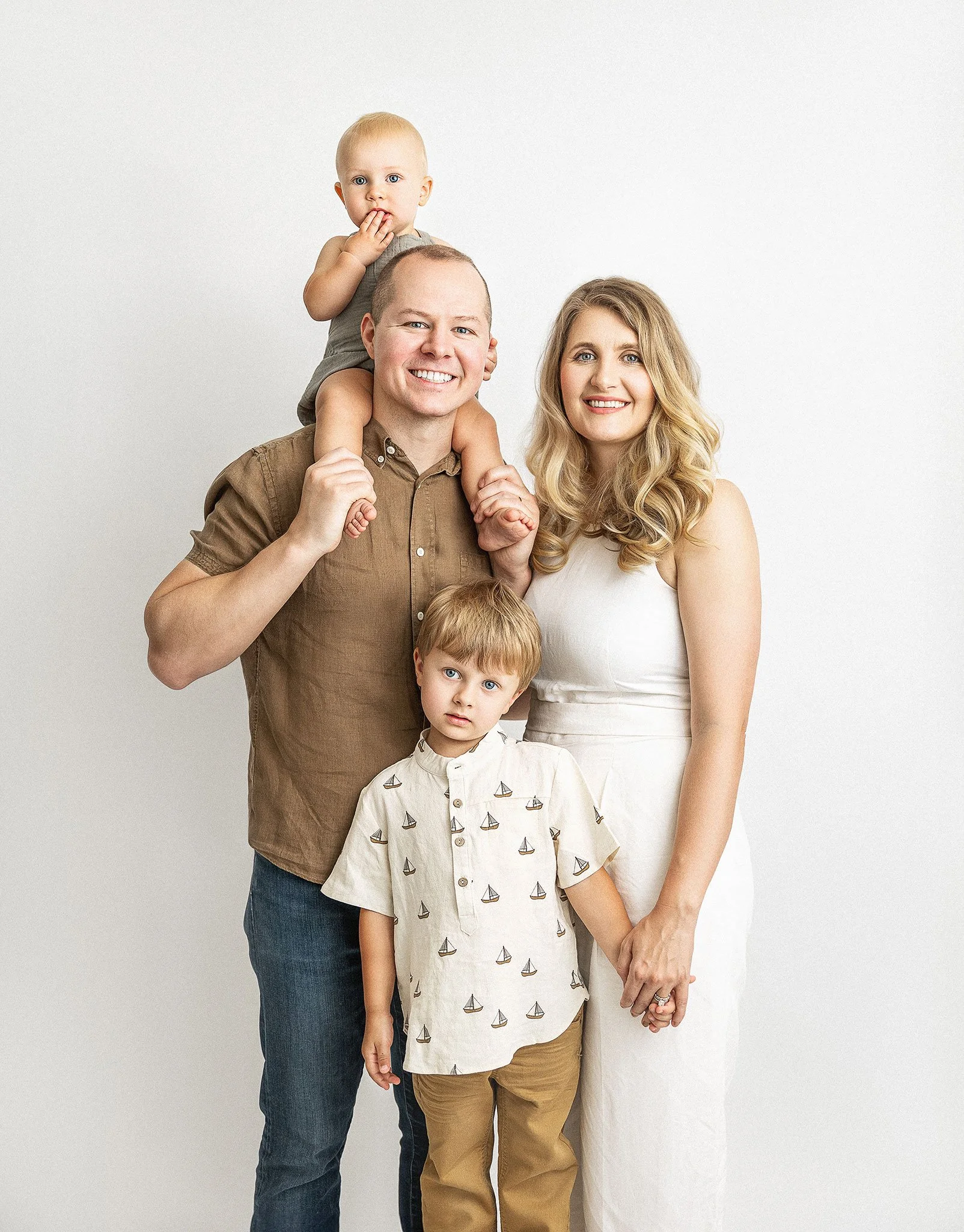 A family portrait with four people standing in front of a plain background. A man with short hair is holding a baby on his shoulders. The woman beside him is smiling, and a young boy stands in front of them, holding her hand. The adults wear casual c