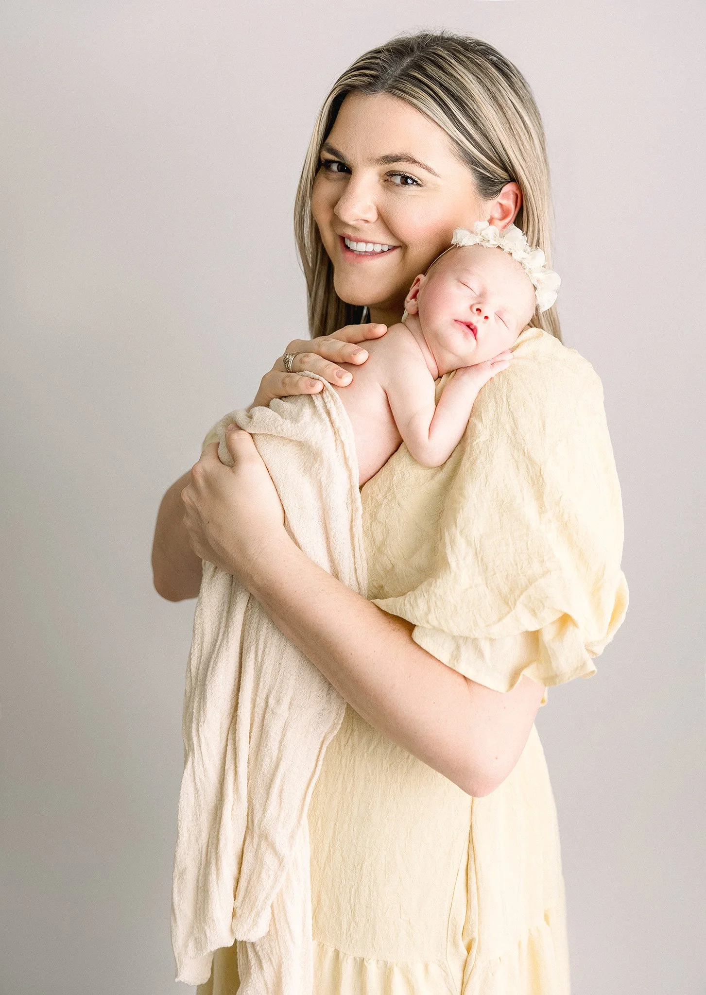 A woman holding a sleeping newborn baby close to her shoulder, with the woman smiling at the camera, both wearing light-colored clothing against a neutral background.