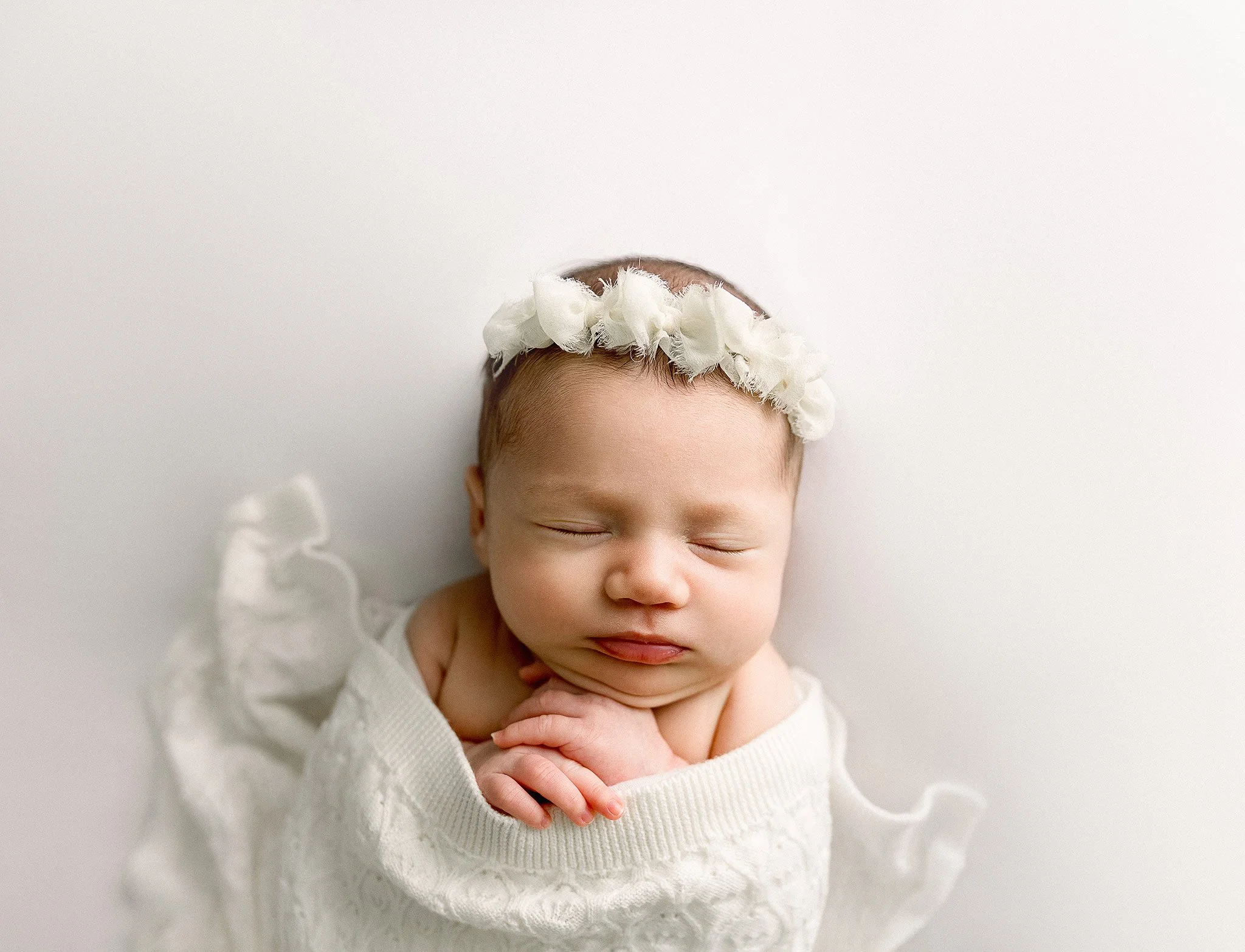 A newborn baby peacefully sleeping, wrapped in a white blanket, wearing a white flower headband, against a plain white background.