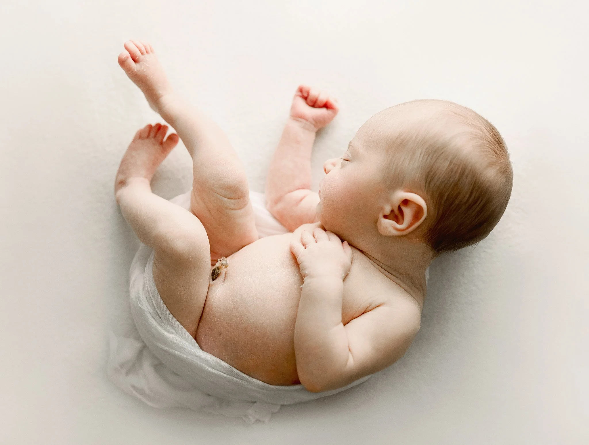 A newborn baby lying on its side on a white surface, with one hand resting on its chest and the other near its face, wearing only a diaper.