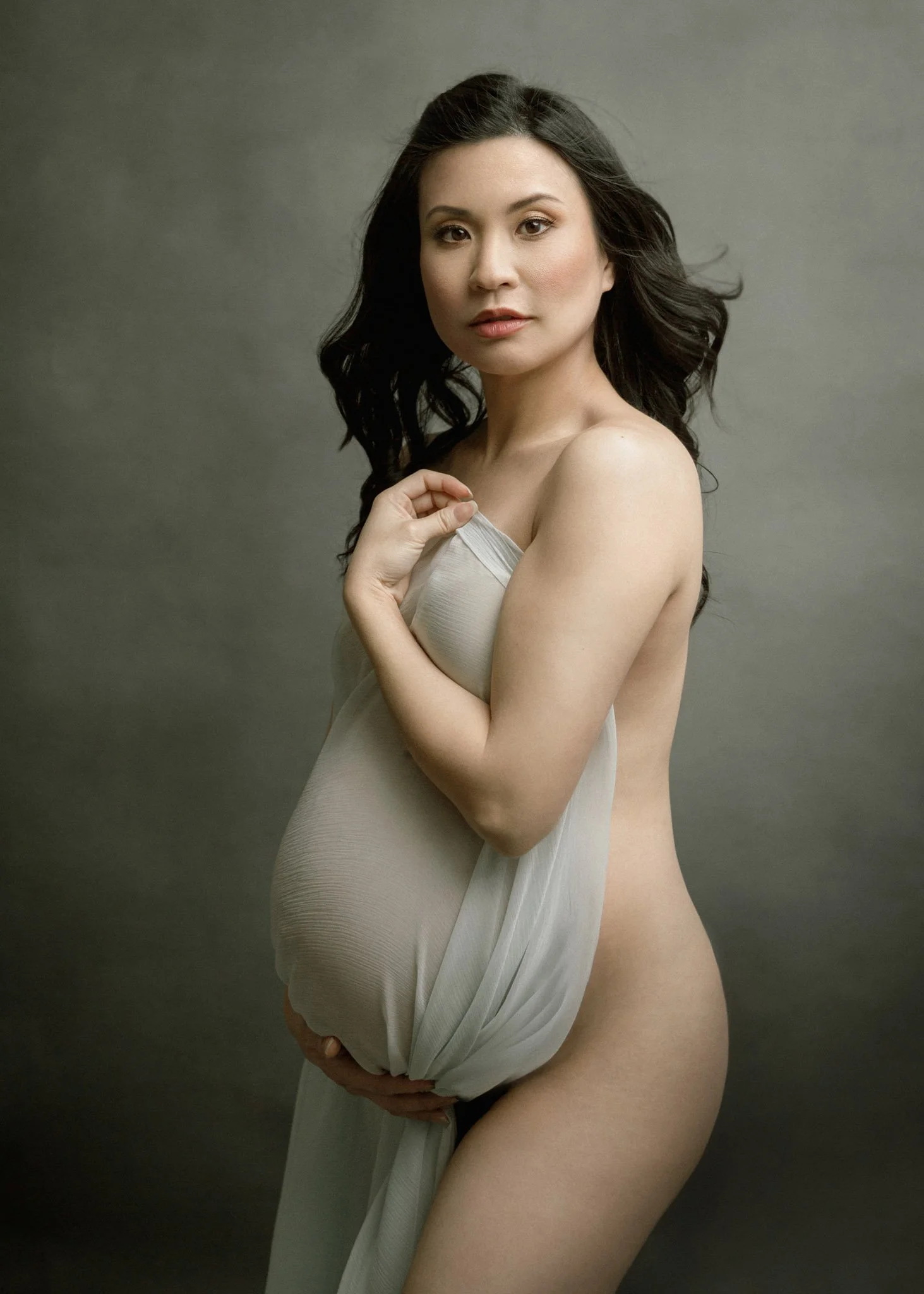 Pregnant woman in profile, draped with light fabric, gazing at camera.