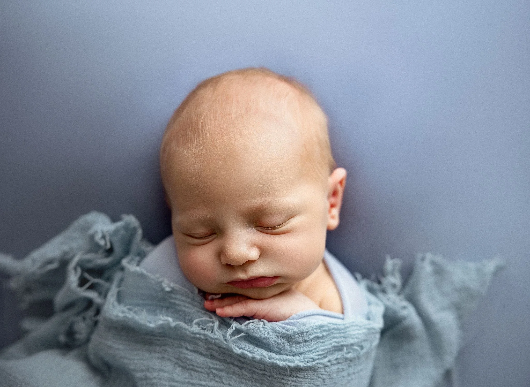 Close-up of a sleeping baby with a peaceful expression, resting on a soft surface, wrapped in a grey blanket.