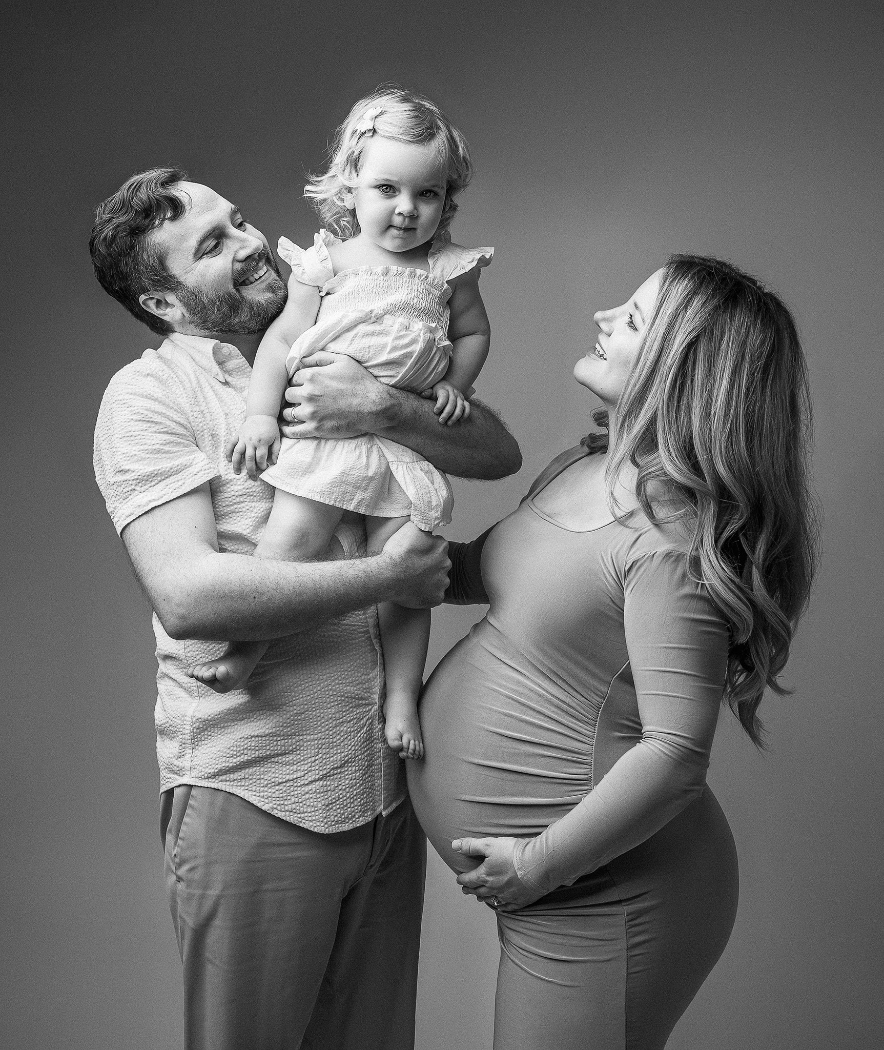 A black and white photo of a smiling family. The man is holding a young child, and the woman, who is pregnant, is looking at them. The child is wearing a dress.