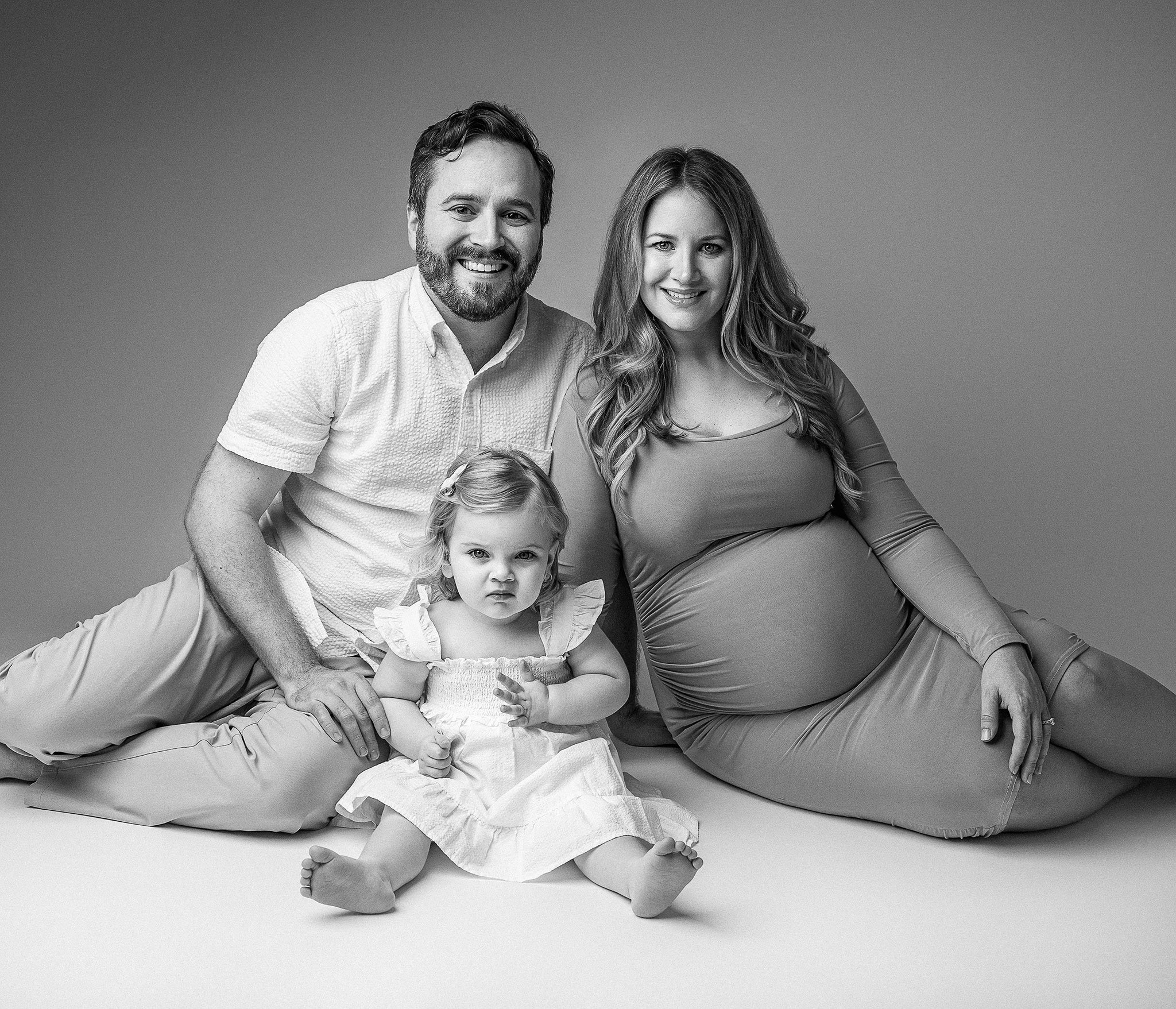 Black and white photo of a smiling family consisting of a pregnant woman in a form-fitting dress, a man wearing a short-sleeved shirt, and a young child in a dress. They are seated and looking at the camera.