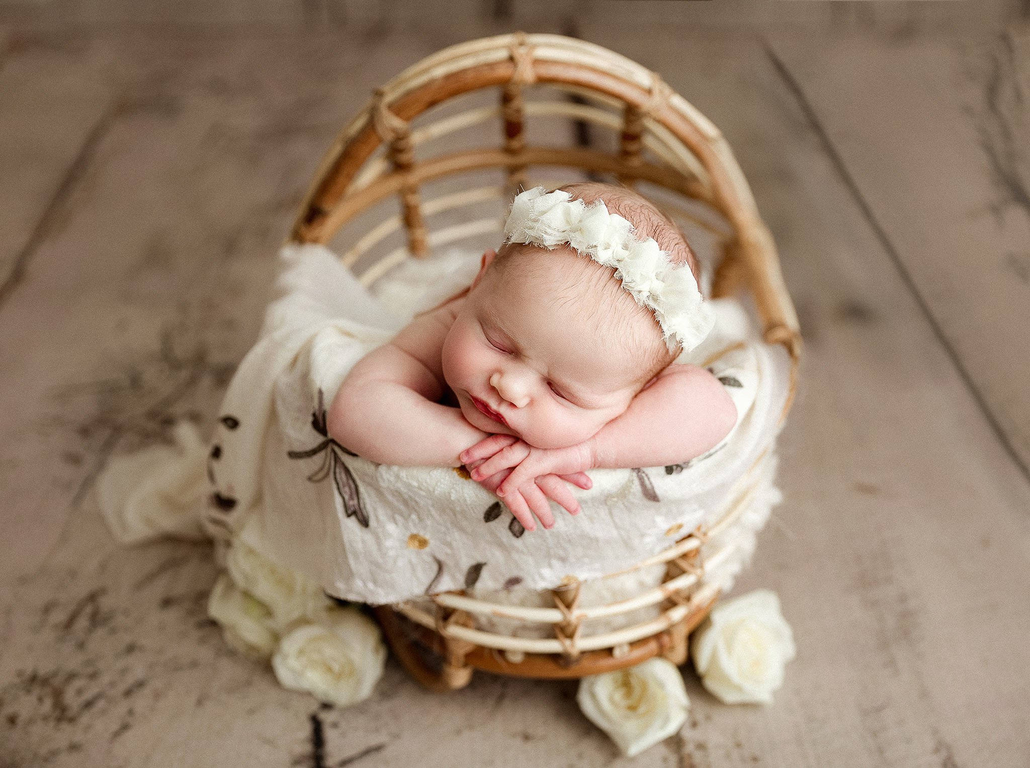 Newborn baby sleeping in a wicker basket, adorned with a white floral headband and wrapped in a soft blanket with flowers.