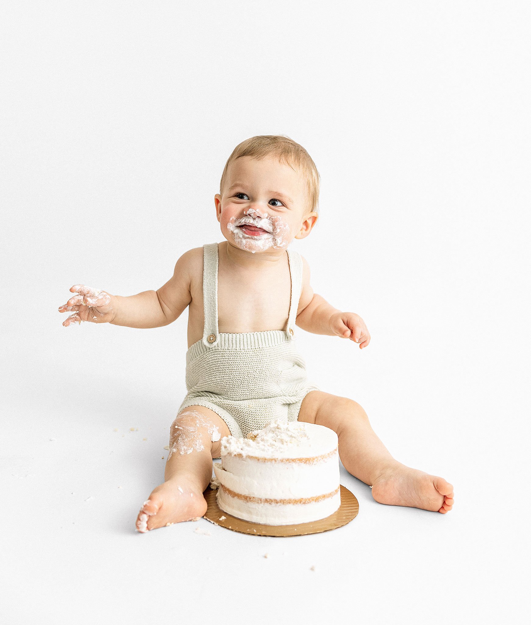 Baby covered in cake sitting on the floor with a small tiered cake.
