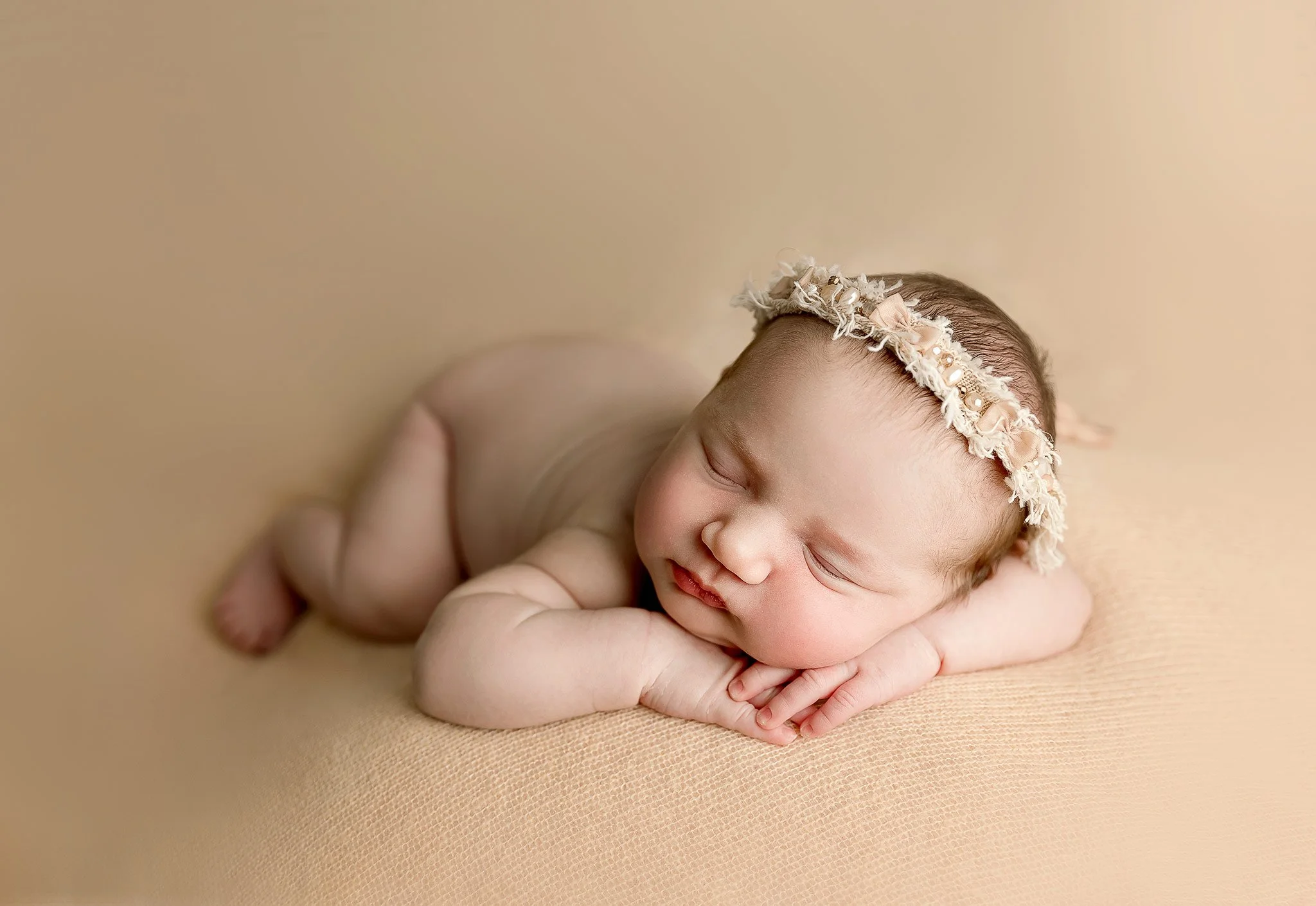 Newborn baby sleeping on beige blanket wearing floral headband.