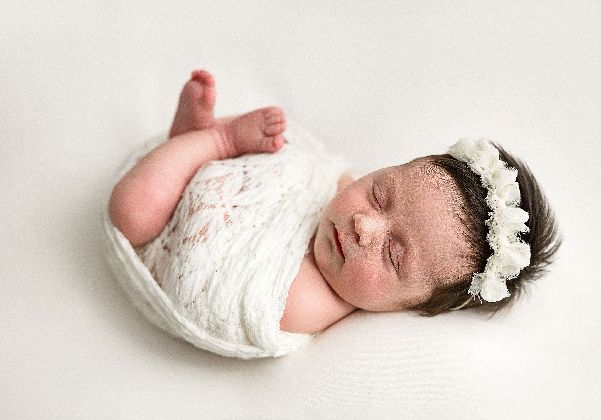 Sleeping newborn wrapped in a white lace blanket, wearing a white floral headband, on a white background - Houston newborn photography