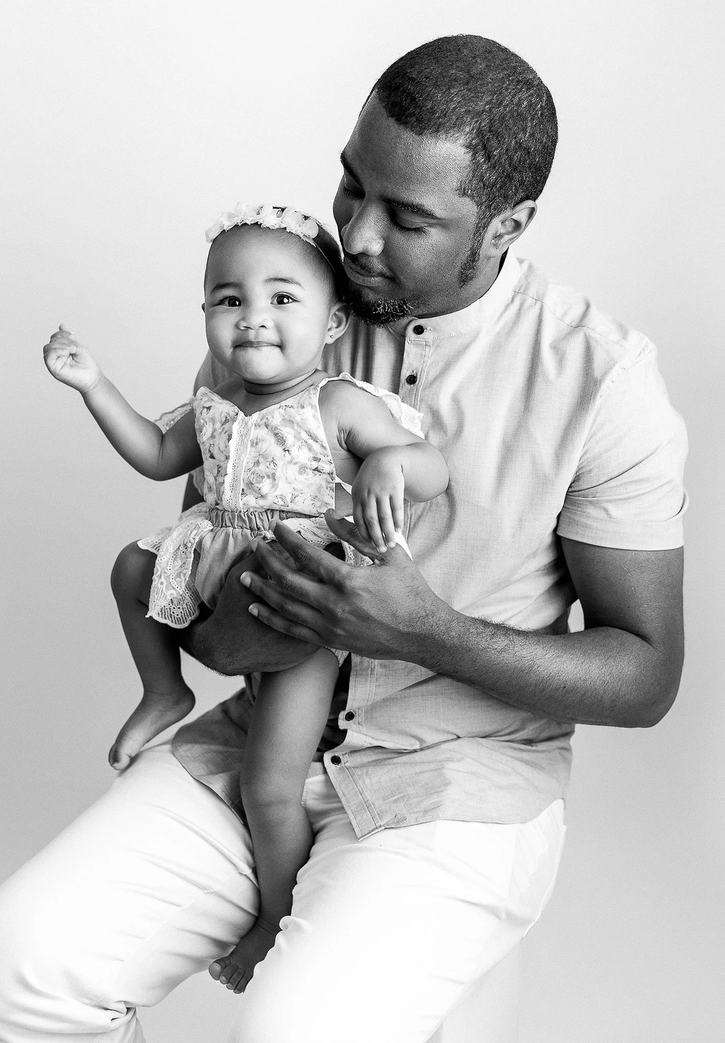 Black and white photo of a father holding his young daughter on his lap. The child is wearing a floral dress and smiling, while the father looks at her lovingly.