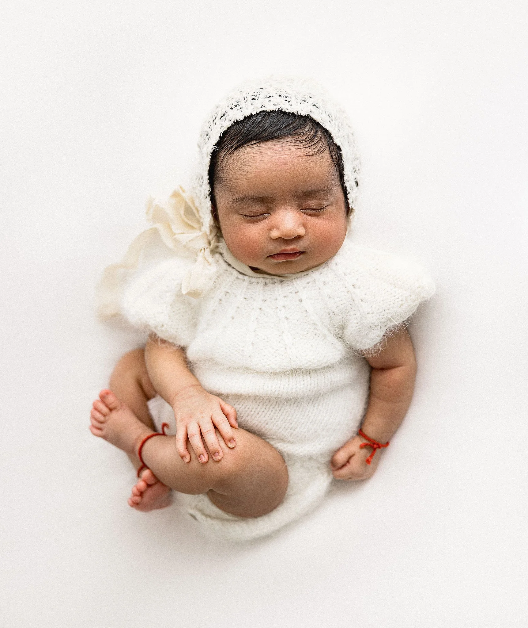 Sleeping baby in white knitted outfit and bonnet on cream background during a newborn session in Houston