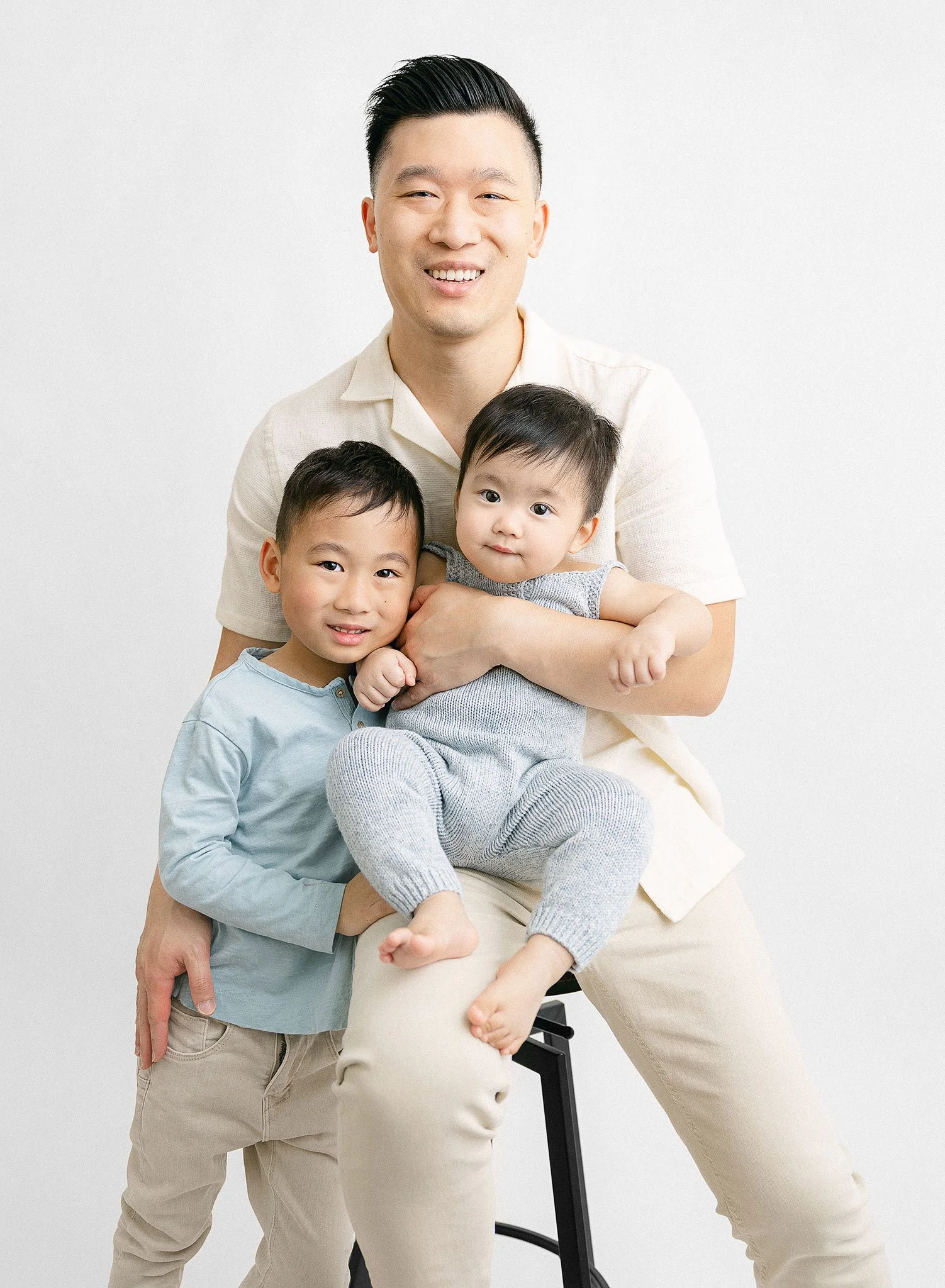 A man sitting on a stool holding a baby, with a young child standing beside them, all smiling against a white background.