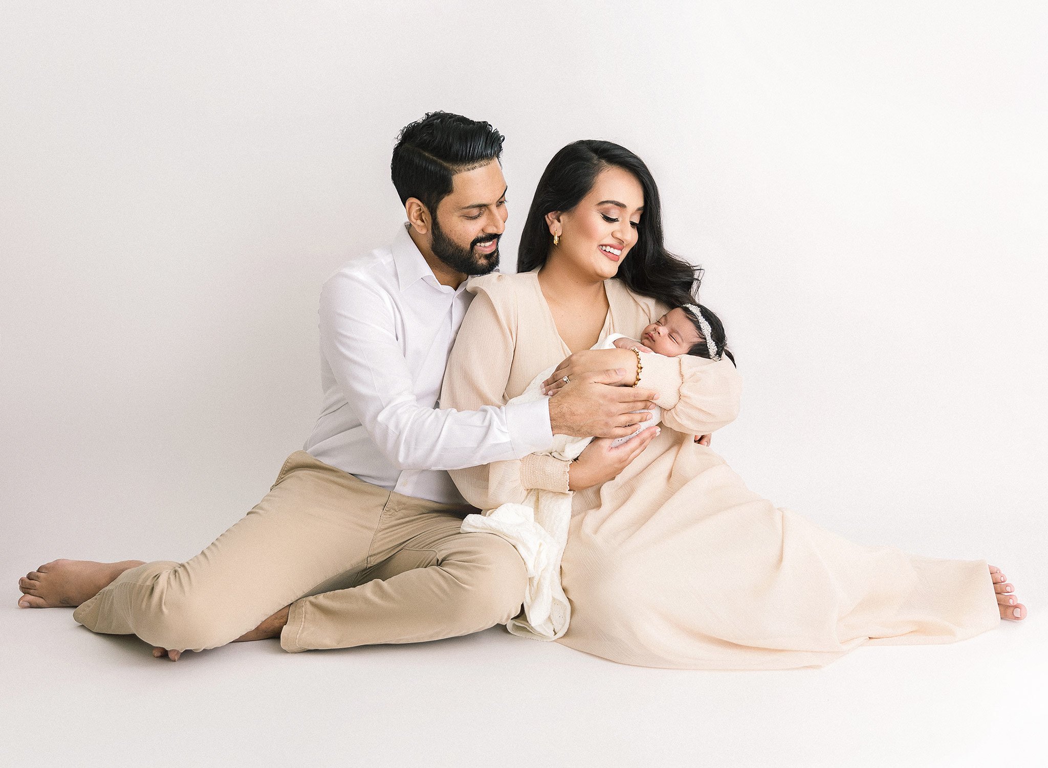 Family portrait of parents sitting and holding a newborn baby against a white background.