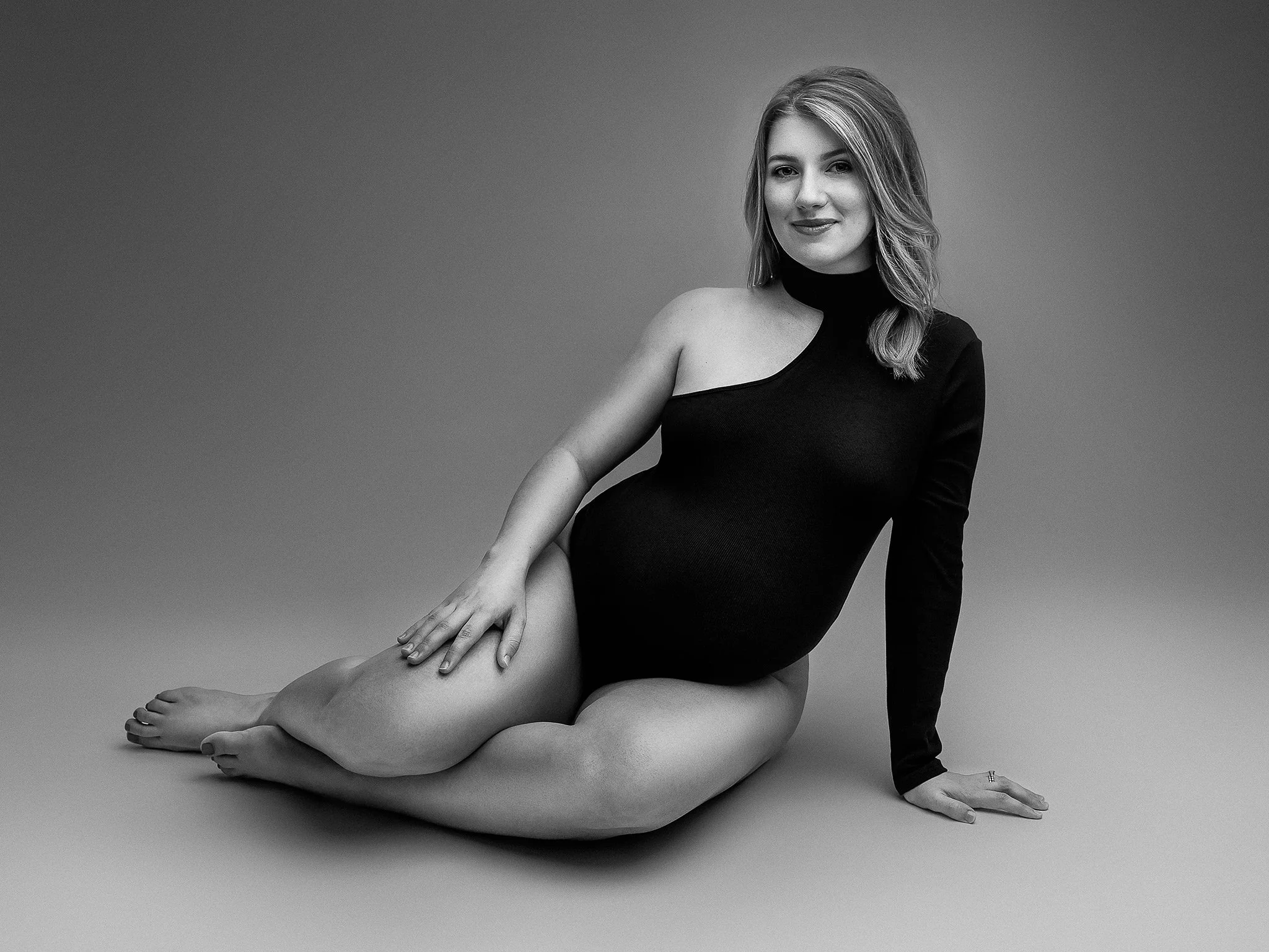 Black and white photo of a pregnant woman sitting on the floor, wearing a one-shoulder black bodysuit.