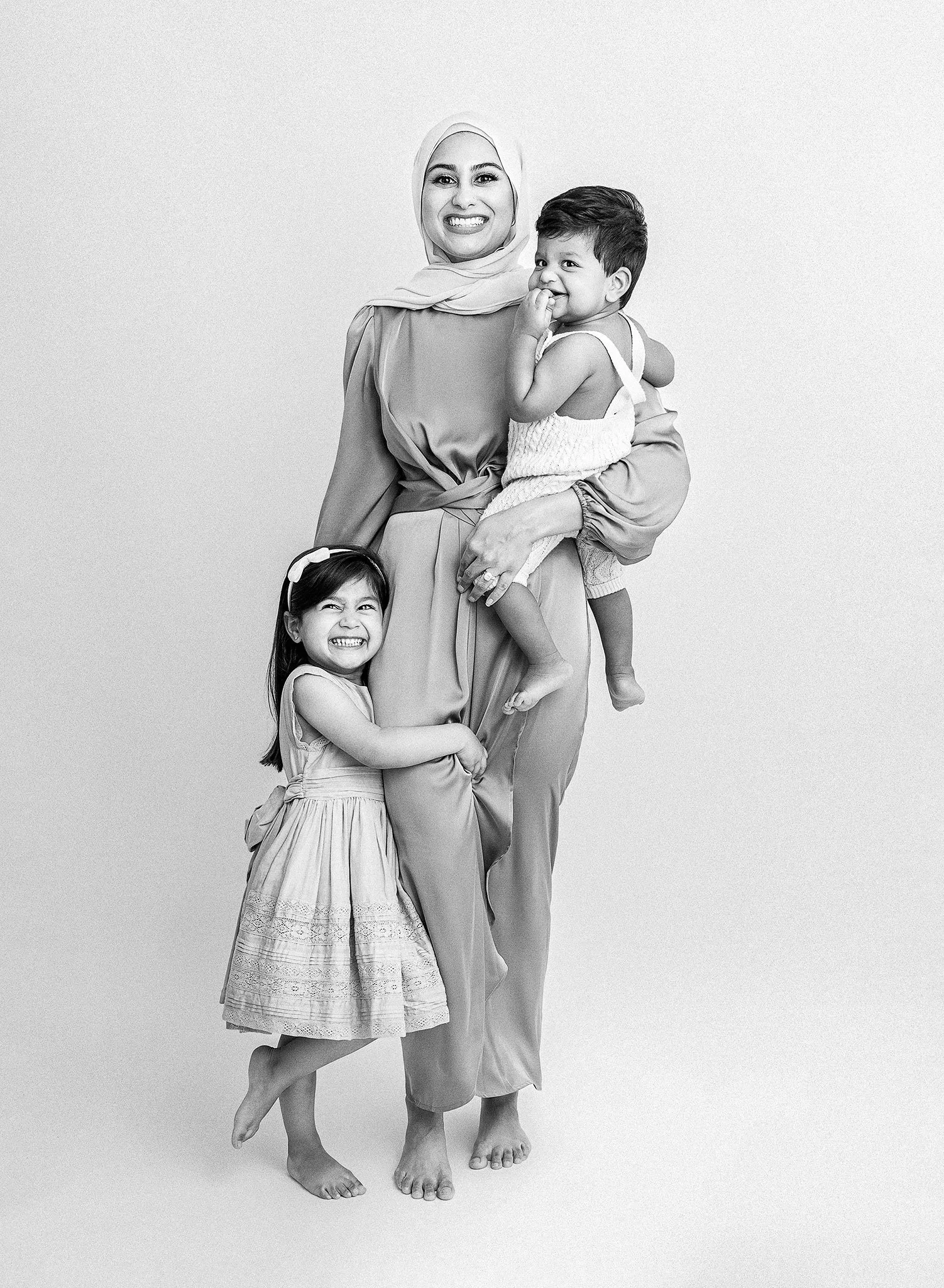 Black and white portrait of a smiling woman in a hijab holding a young child and standing next to a smiling girl, all in light clothing.