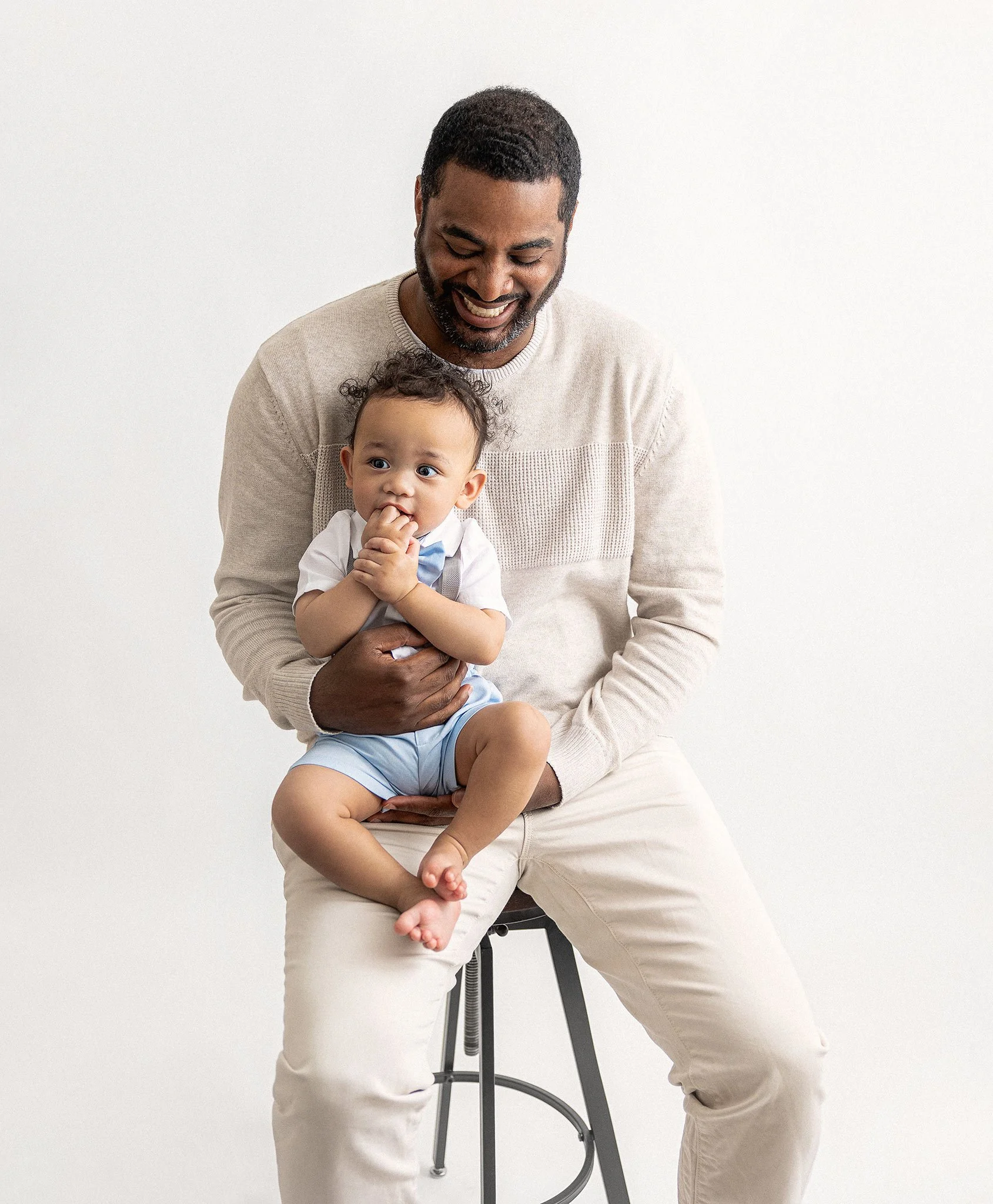 Smiling man holding a baby on his lap, both sitting on a stool against a white background.