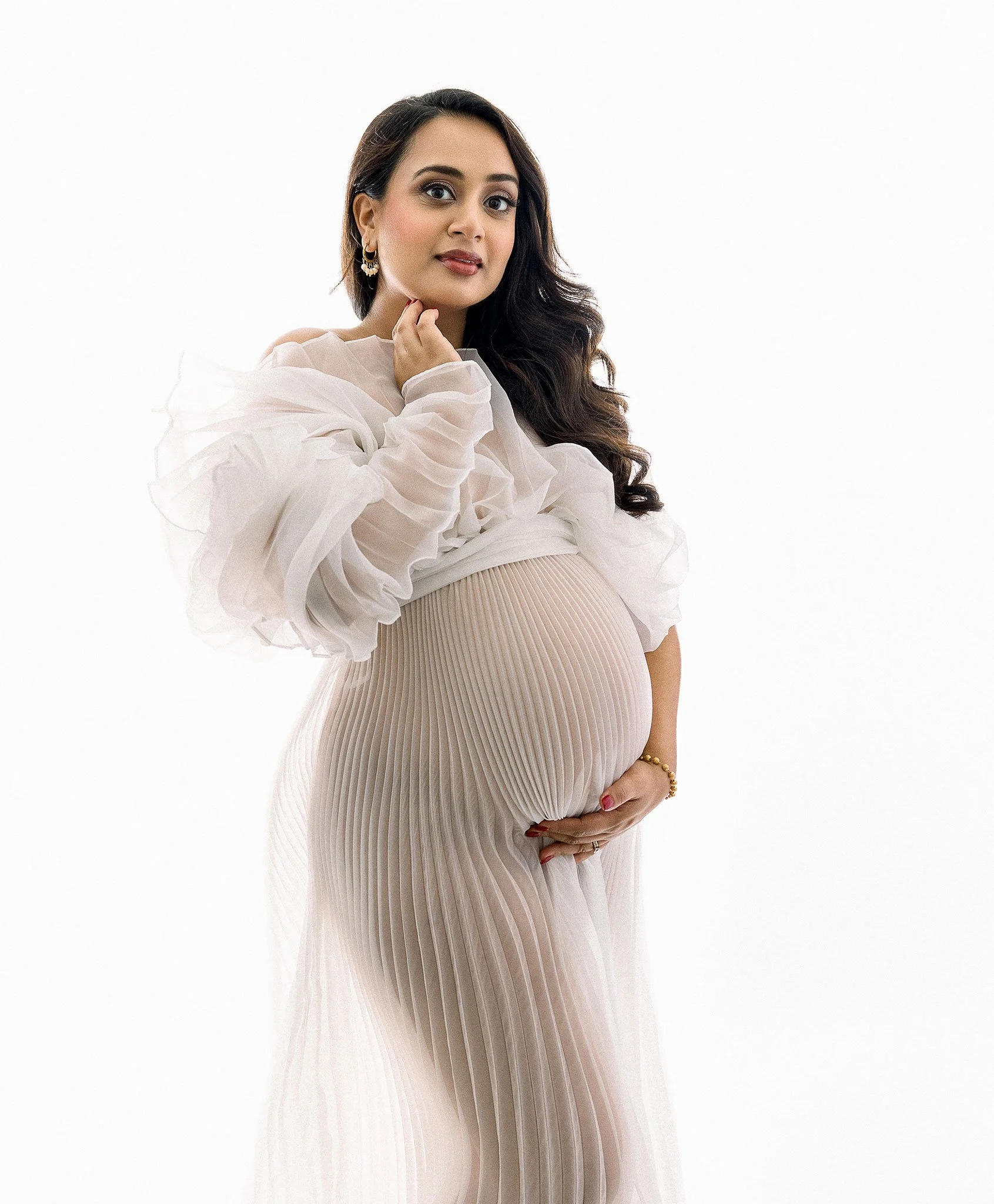 Pregnant woman in white pleated dress, holding belly, with long hair and gentle expression against plain background.
