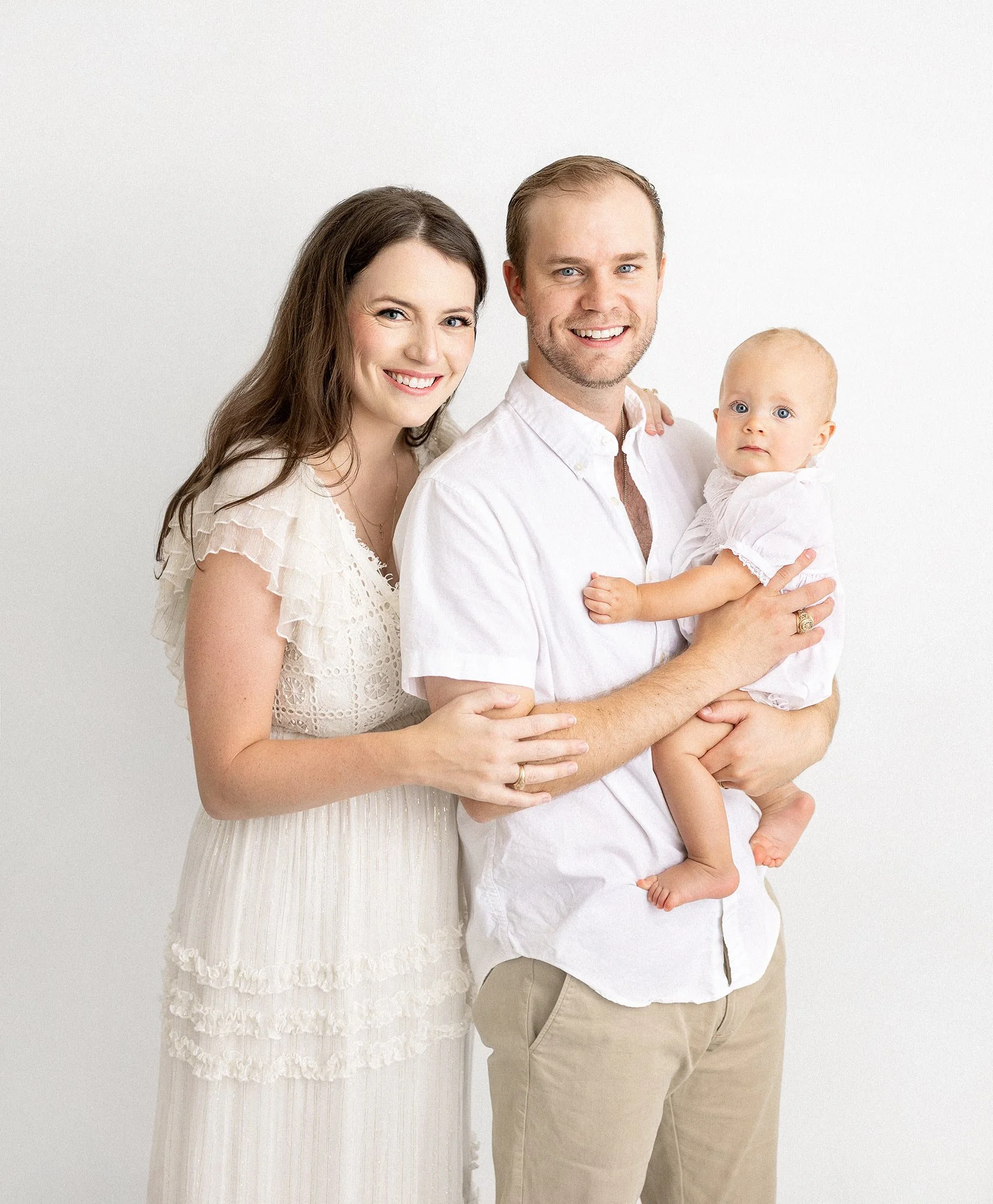 A smiling couple with a baby, posing in a light, airy setting. The woman is wearing a white lace dress, the man is in a white shirt and beige pants, and the baby is dressed in white.