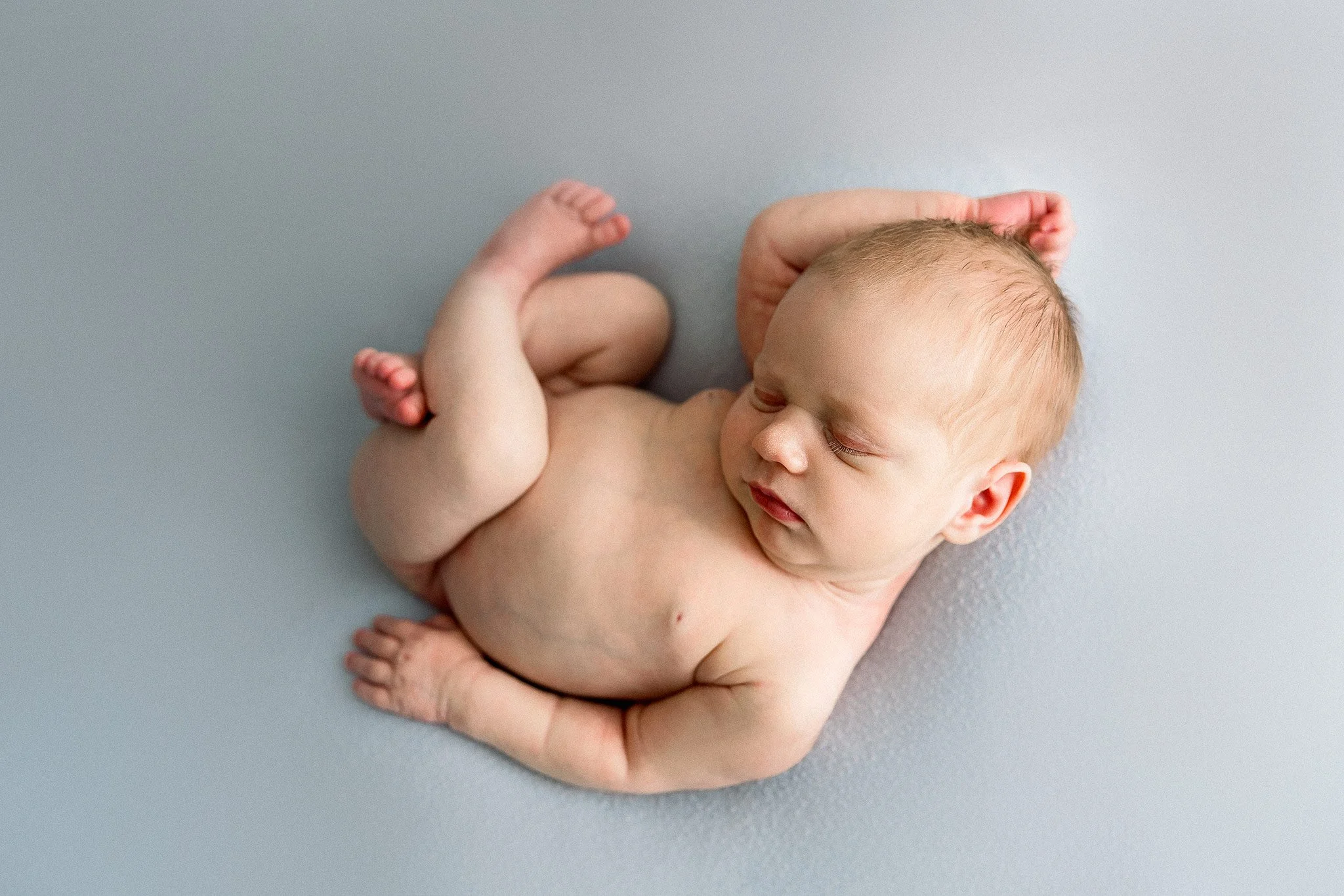 A newborn baby sleeping on a light blue surface with arms and legs curled up.