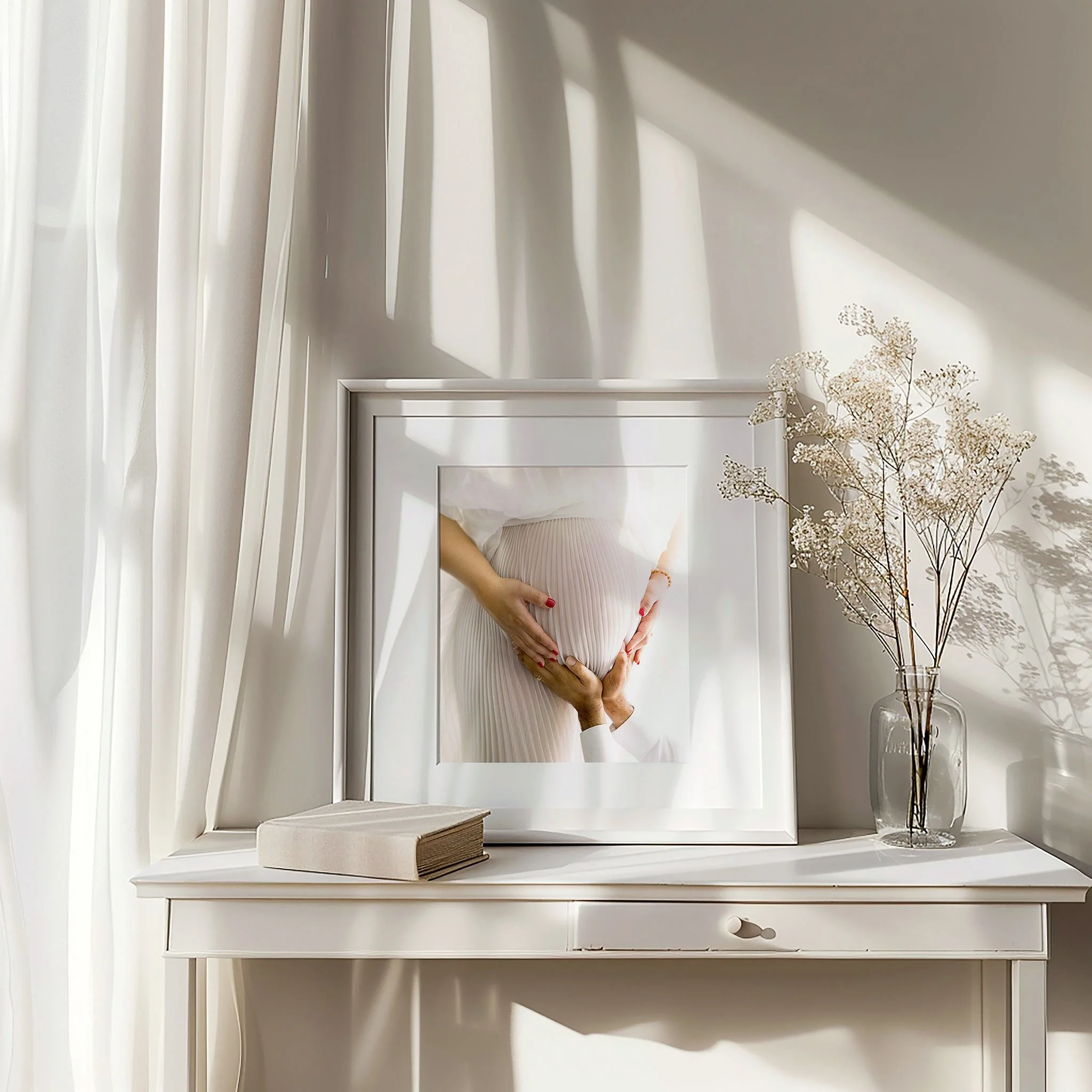 A white dresser with a book and a glass vase filled with dried white flowers placed on top. White vertical blinds cover a window and sunlight casts shadows on the wall.