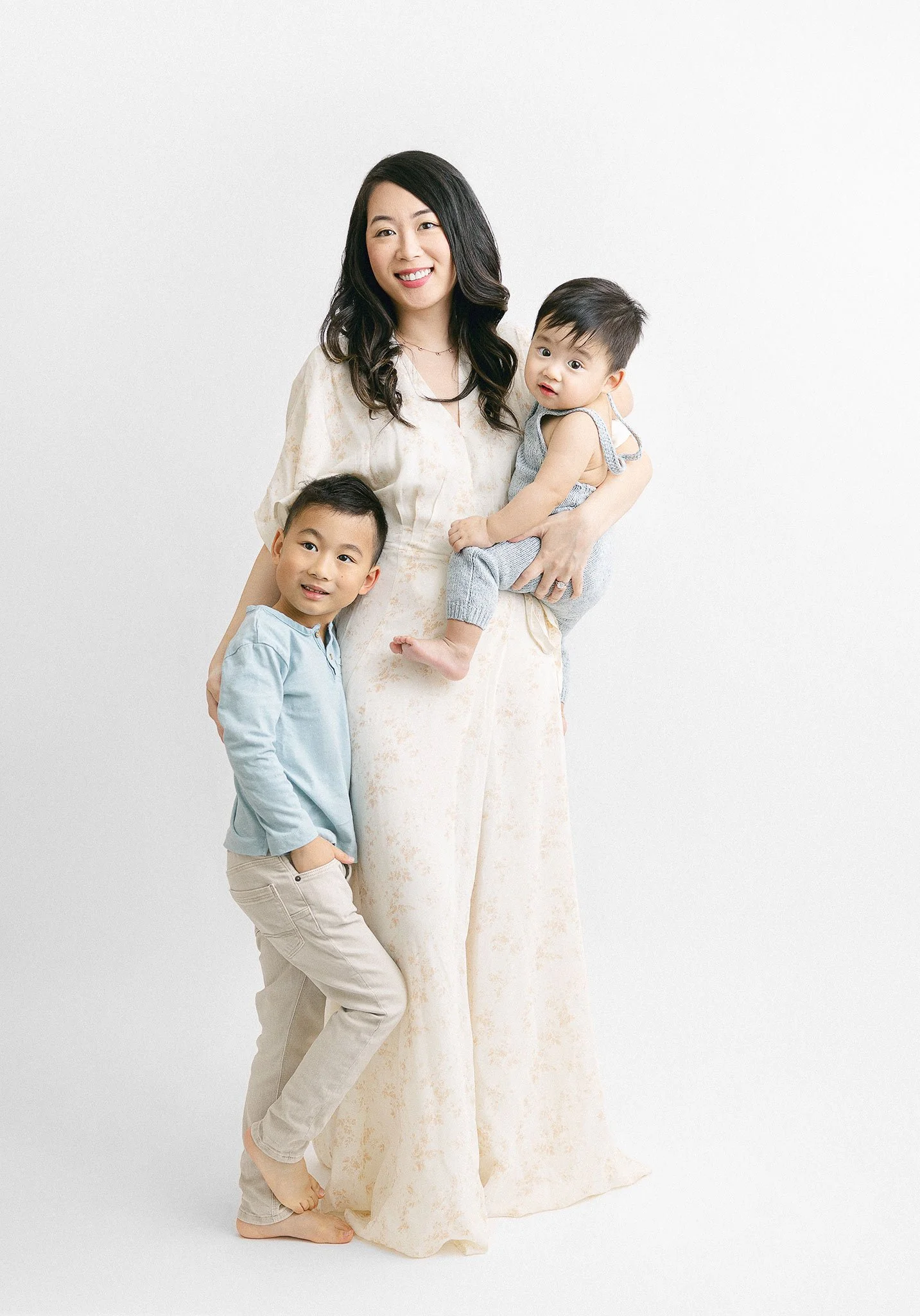 A woman in a white dress holding a baby, with a young boy standing next to her in a blue shirt and beige pants. They are posing against a white background.