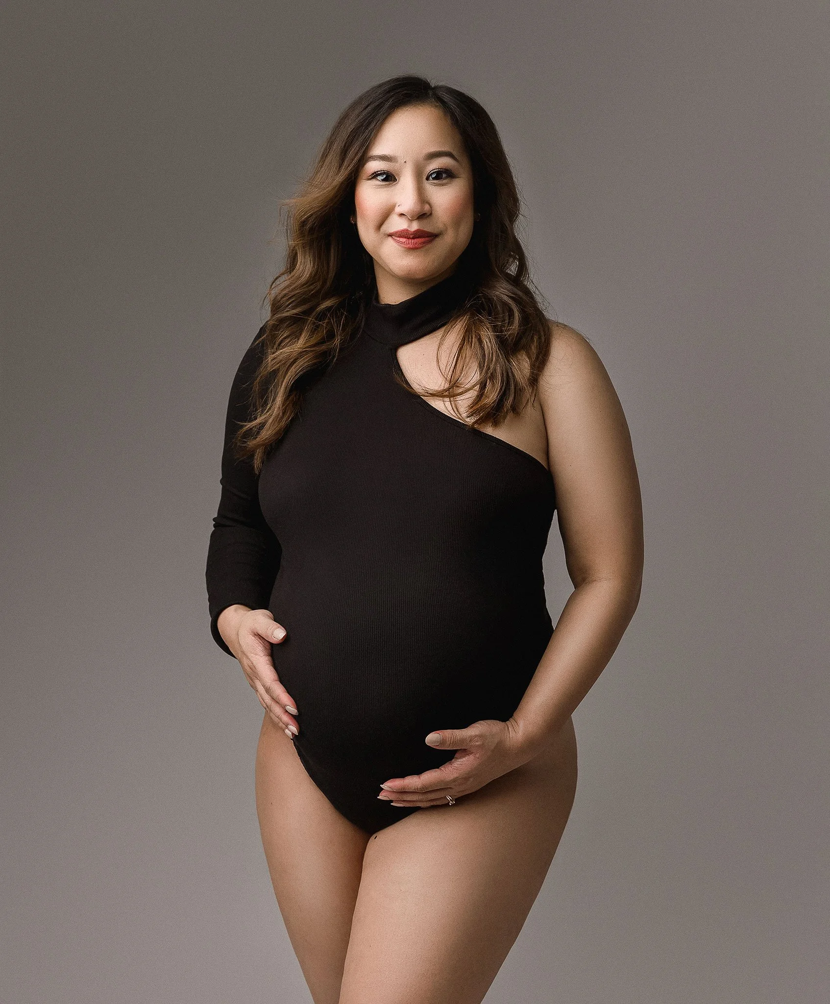 Pregnant woman in a black one-shoulder bodysuit, holding her belly, against a neutral background.