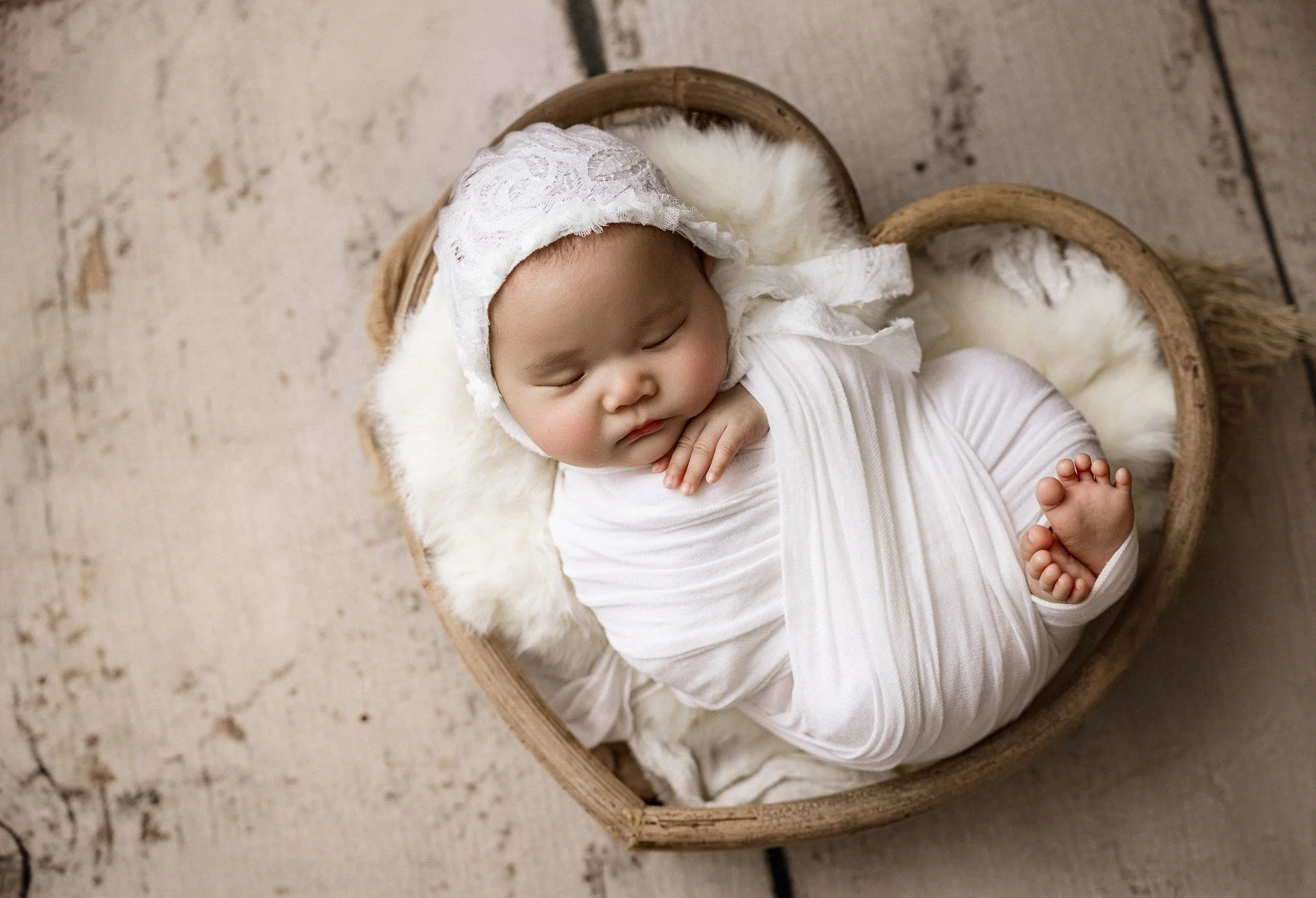A sleeping baby girl curled up in a heart-shaped wooden basket, wrapped in white swaddle and wearing a lacy white bonnet, on a wooden floor.