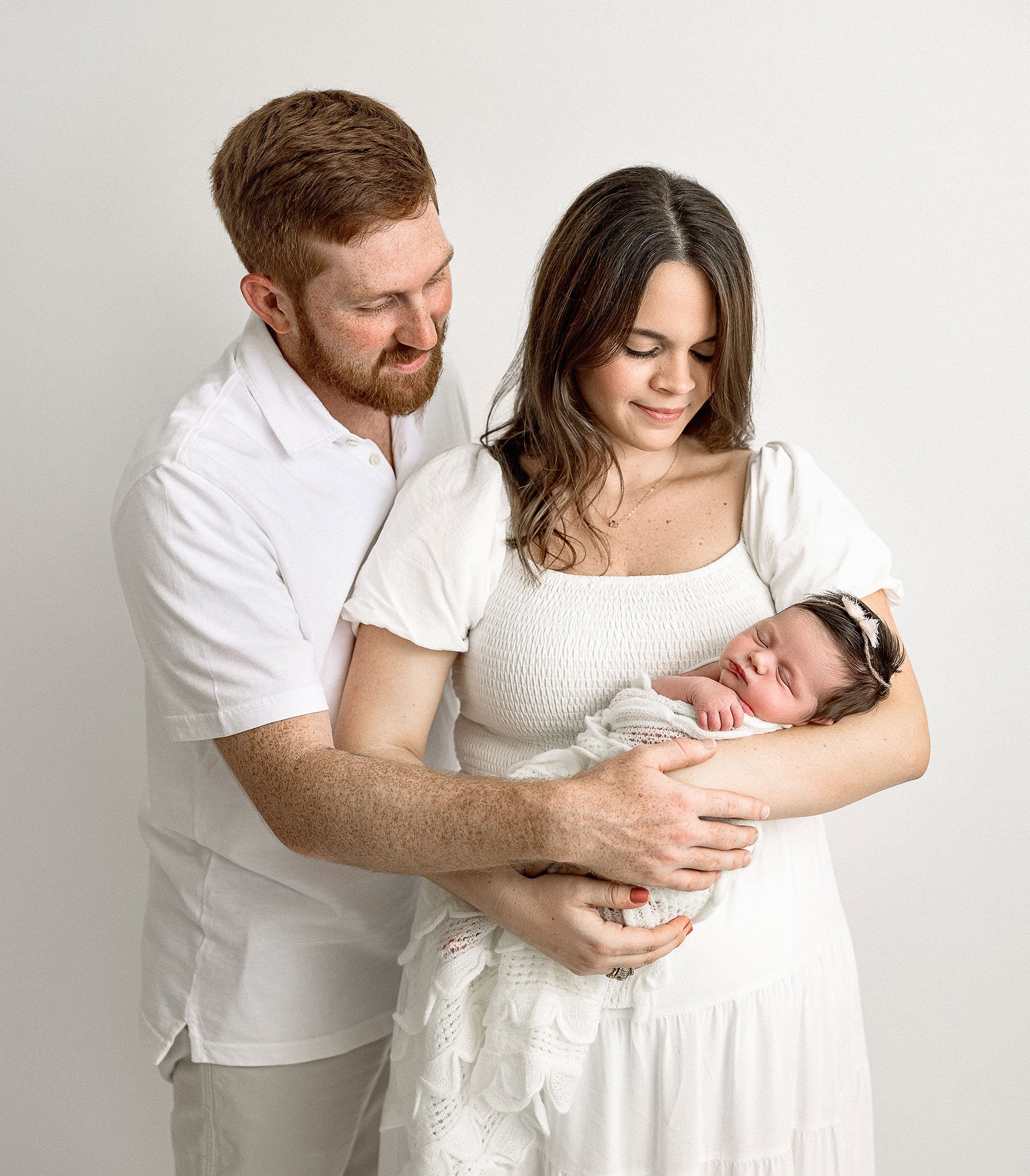 A couple in white clothing lovingly holding a sleeping newborn wrapped in a blanket against a plain background.