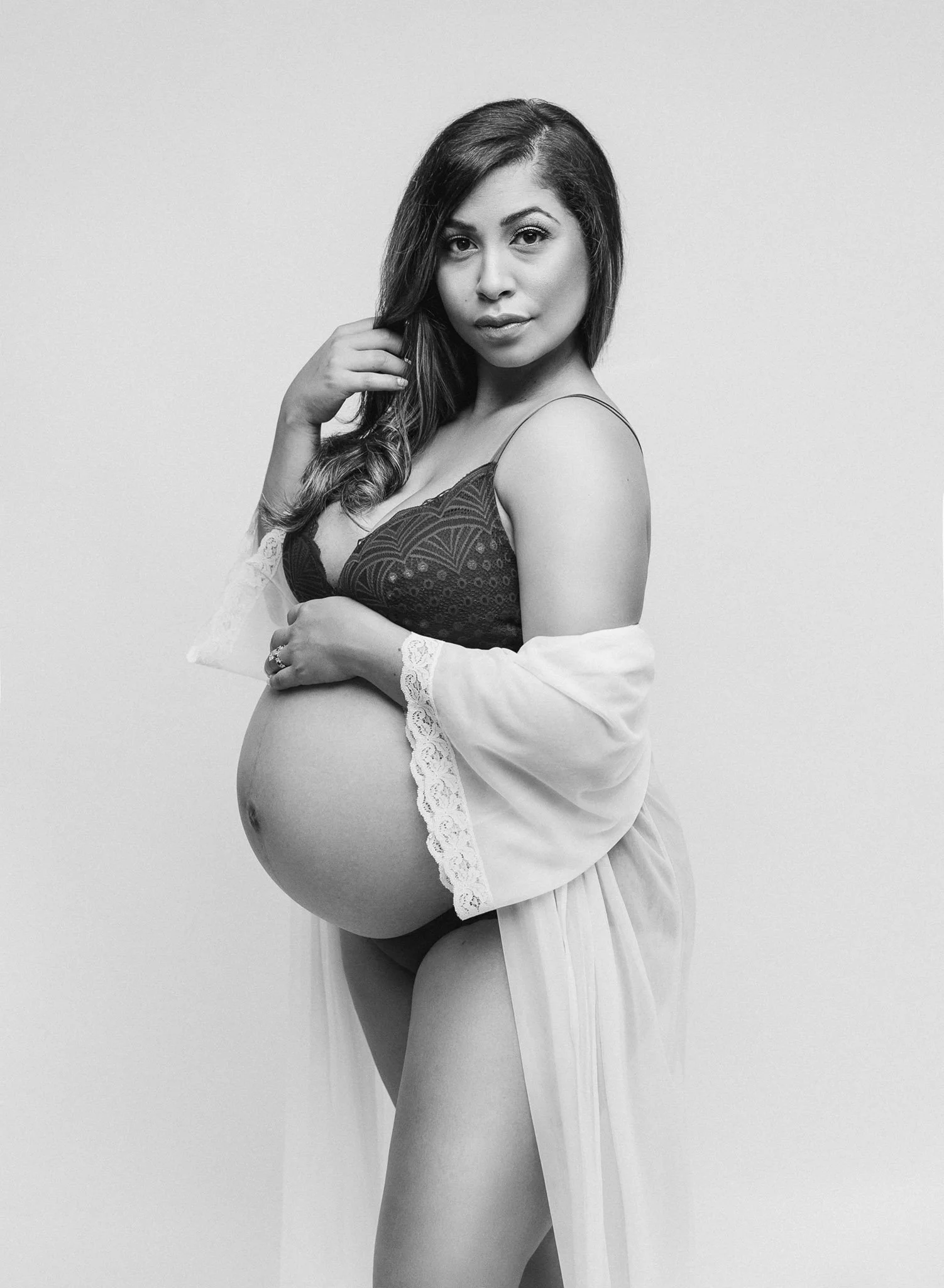 Pregnant woman in lingerie and robe holding her belly in a maternity photoshoot.