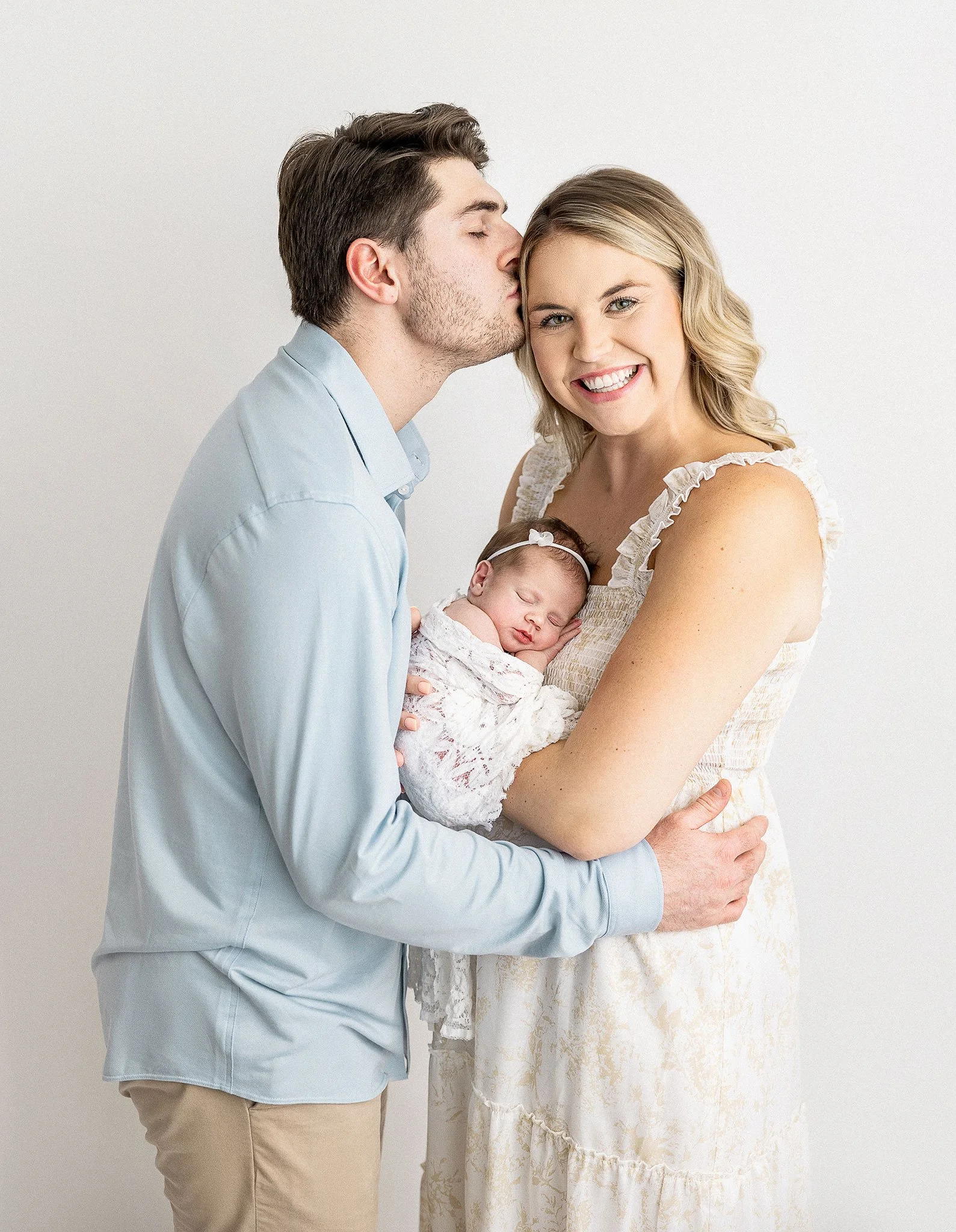 A smiling woman holding a sleeping newborn wrapped in a lace blanket, while a man kisses her forehead.