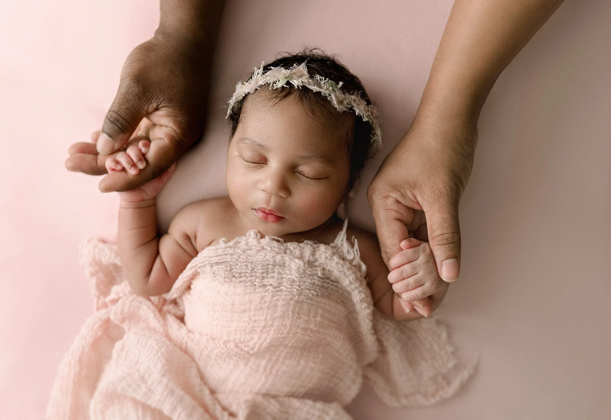 A sleeping baby girl held gently by an adult, with her eyes closed and wearing a pink headband and a matching pink blanket, on a soft pink background.